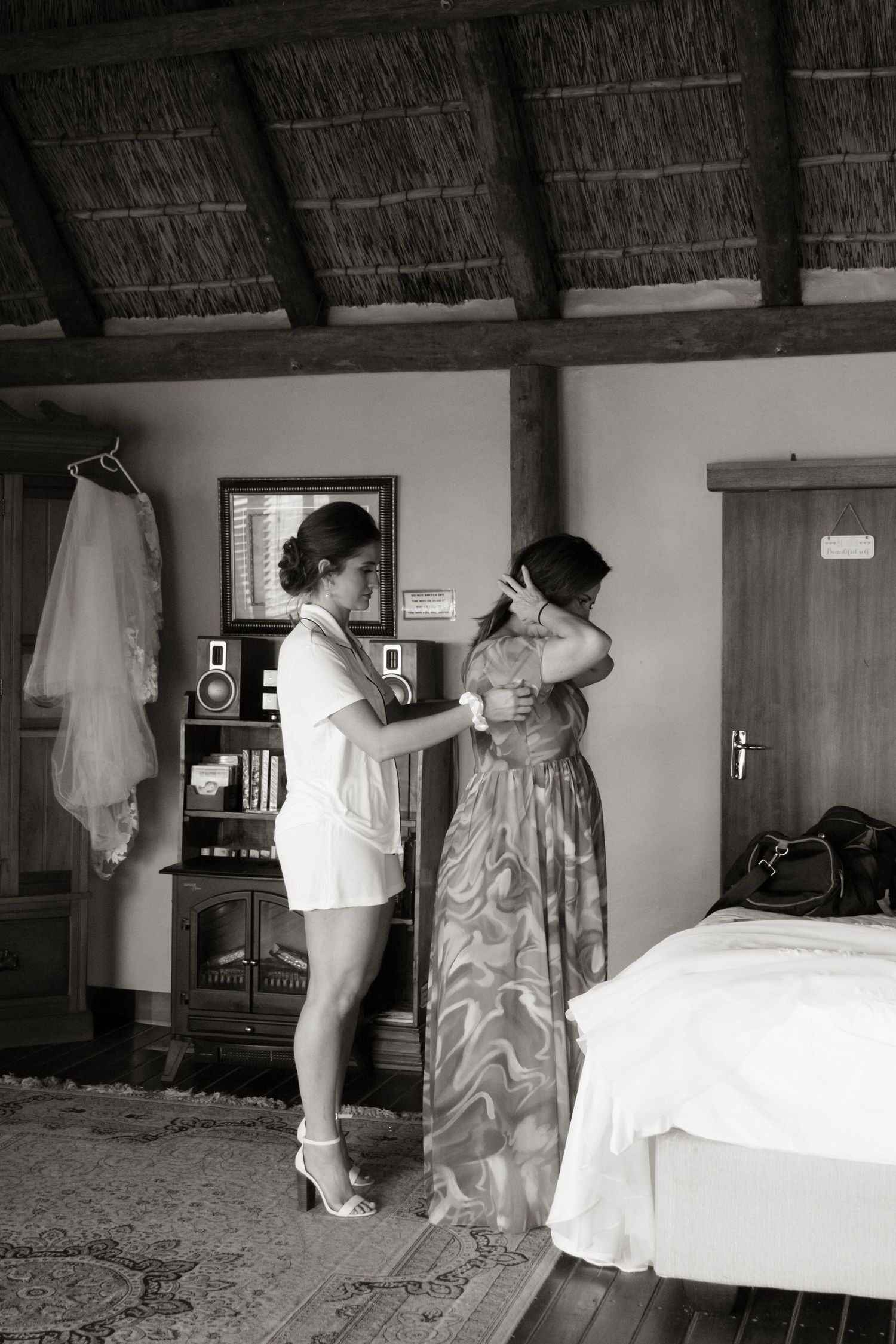 Black and white photo of two women getting ready for a wedding in a rustic thatched room.