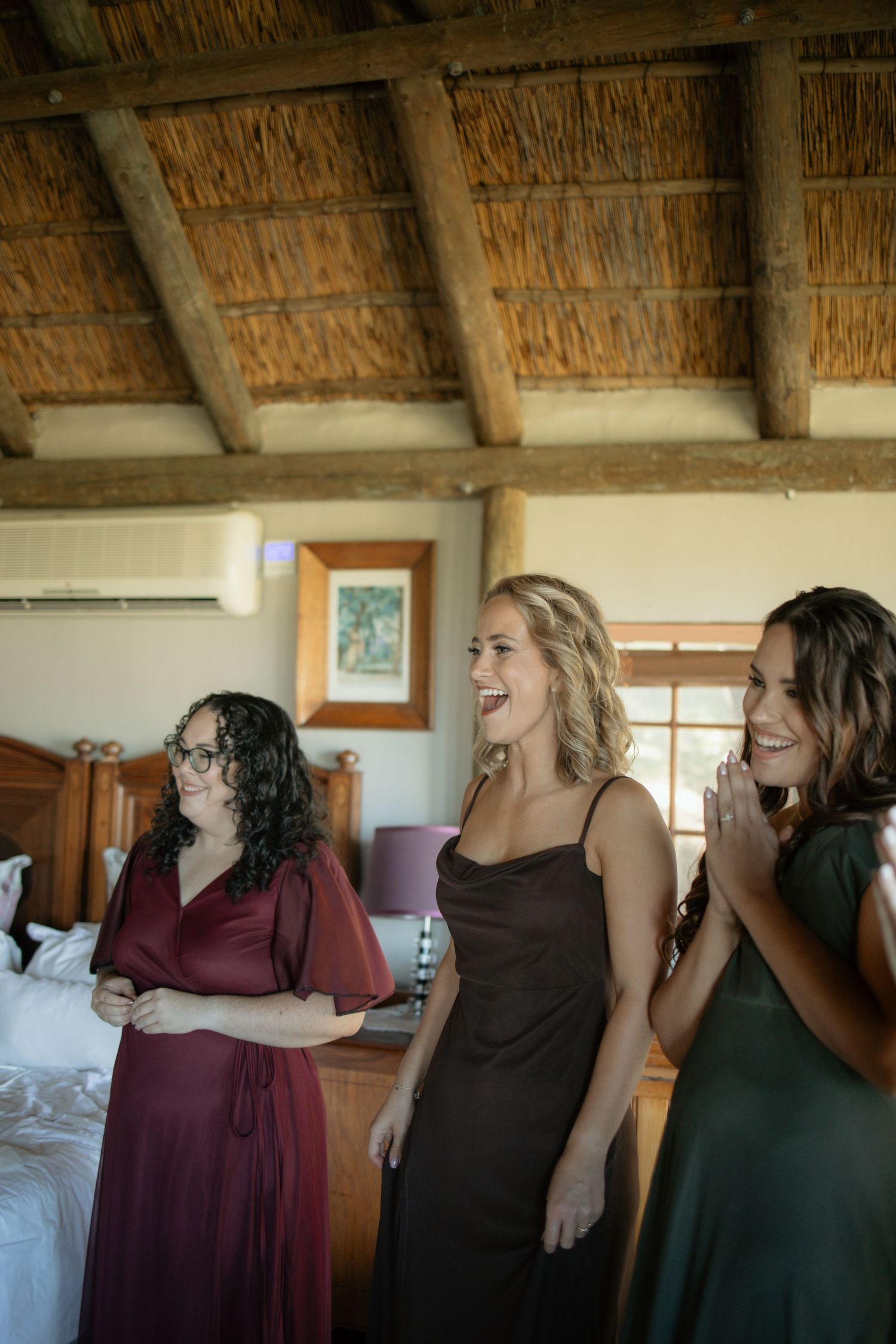 Three bridesmaids in colorful dresses laughing joyfully in a thatched-roof room during wedding preparations.