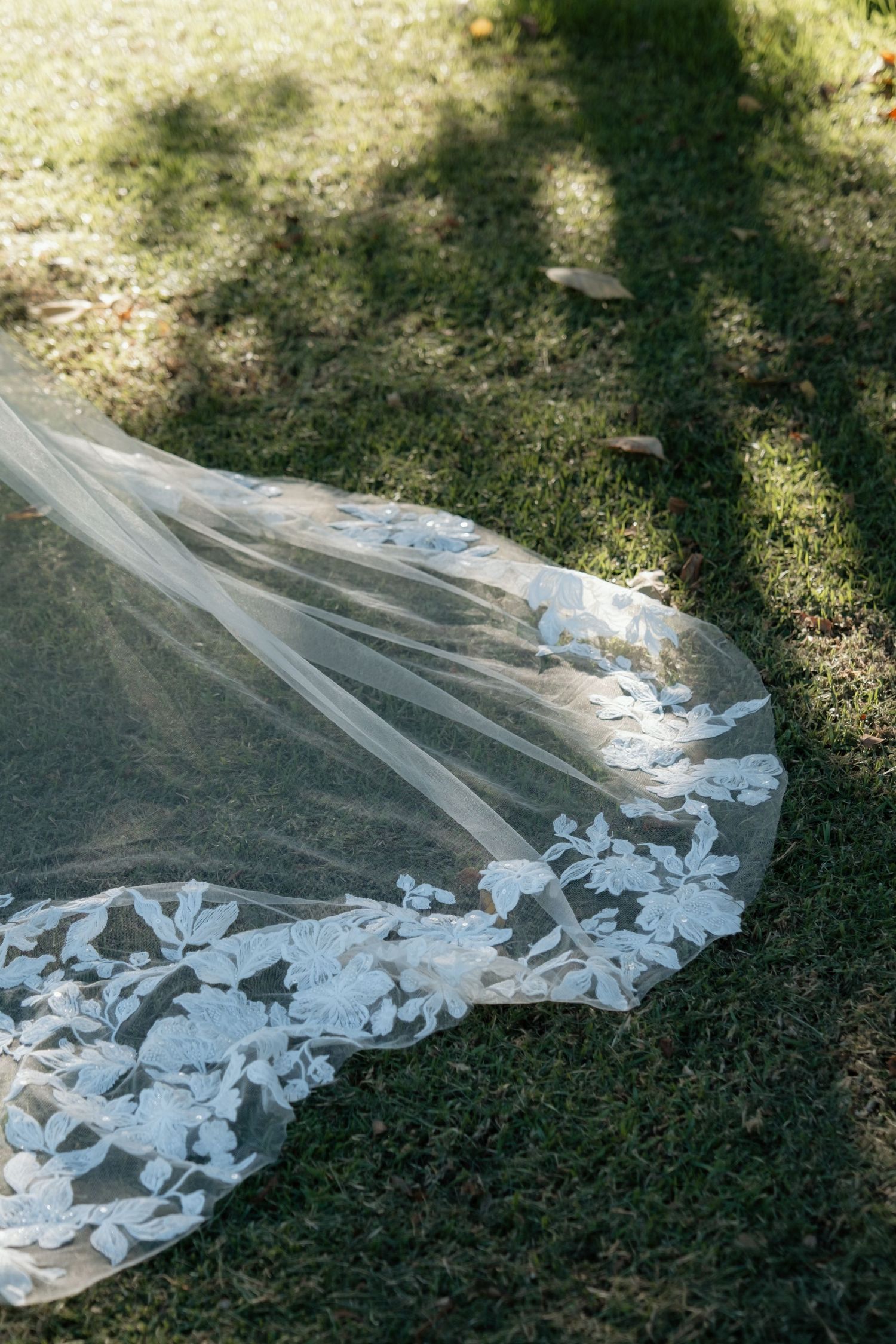 Elegant bridal veil with floral lace trim spread across green grass in dappled sunlight.