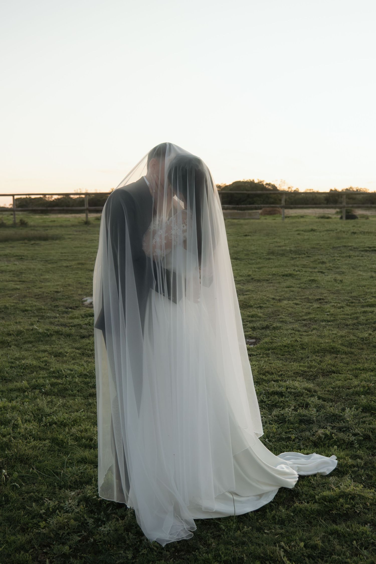 Bride and groom share a kiss beneath a flowing white veil in a green field at dusk.