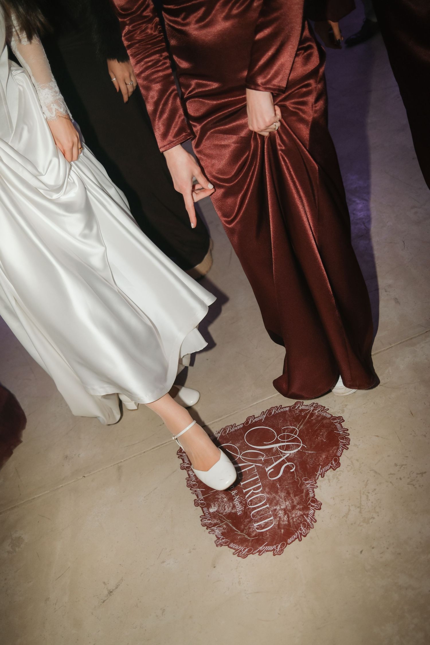 Bride in white dress and heels standing beside woman in burgundy satin gown on decorated wedding venue floor.