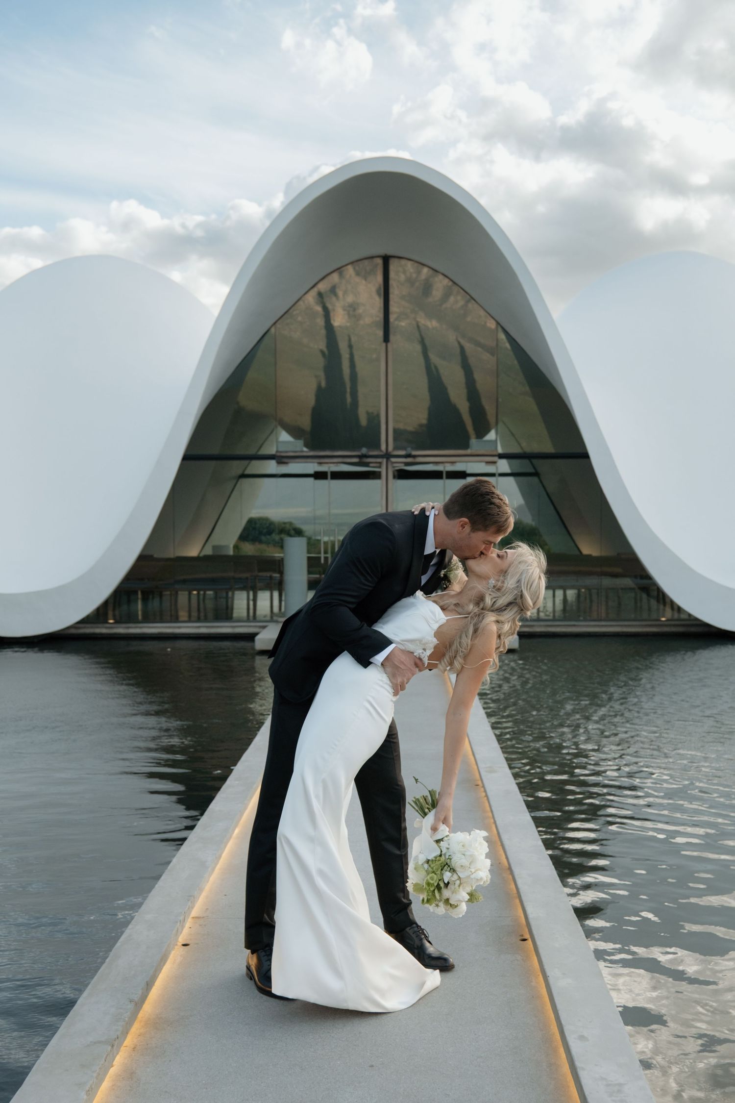 A romantic wedding photo showing a couple dipping and kissing in front of the modern BOSJES Chapel