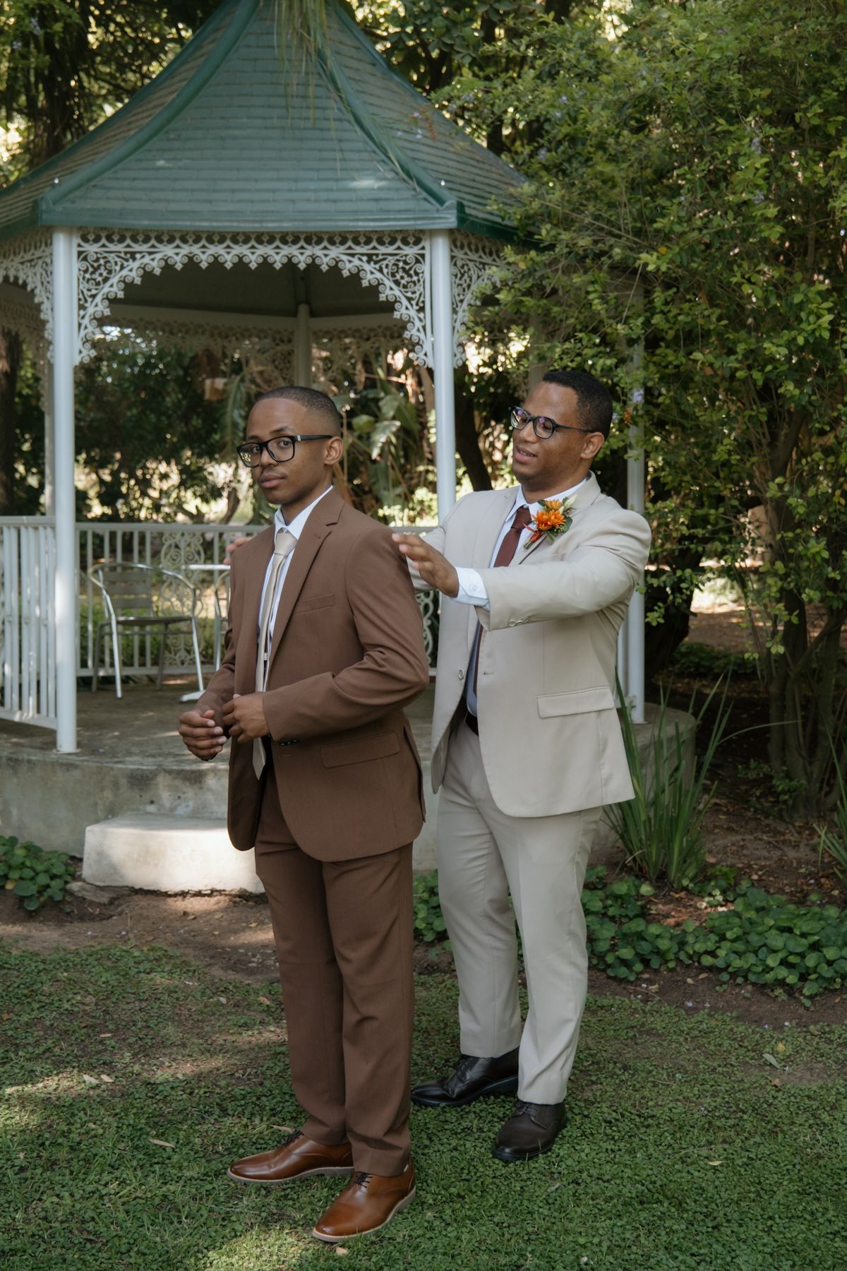 Two grooms in brown and beige suits adjust boutonnieres near a white gazebo in a lush garden wedding setting.