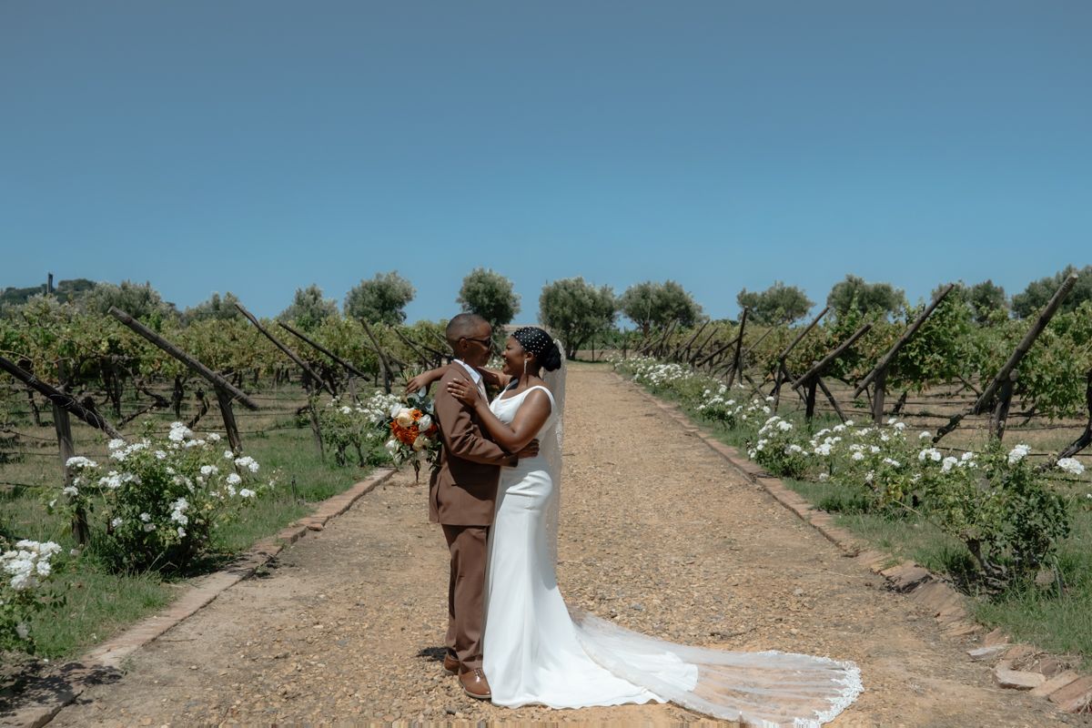 Bride and groom sharing a romantic moment in a vineyard aisle, surrounded by lush green vines under a clear blue sky.