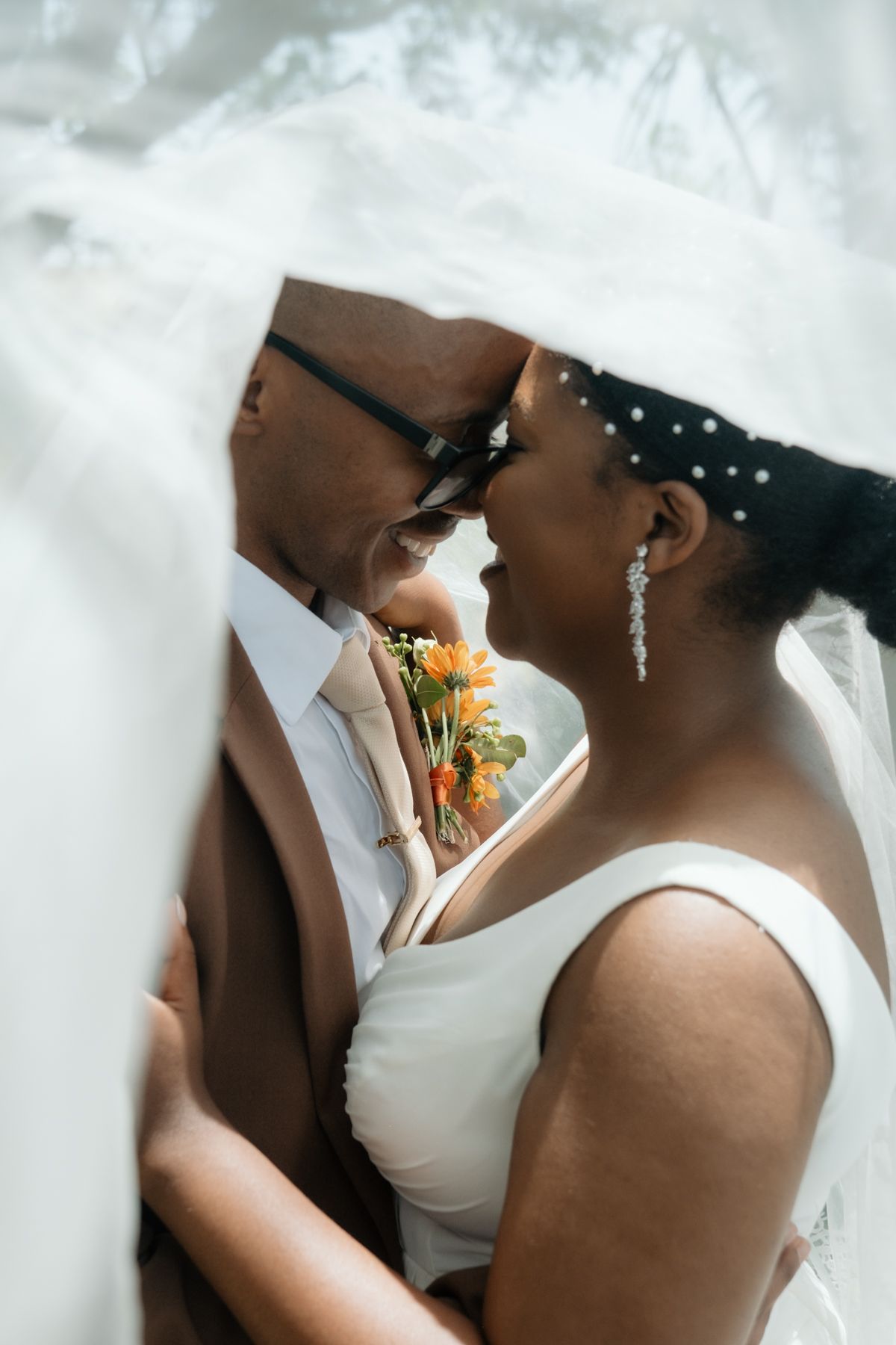 Black bride and groom share intimate forehead touch under wedding veil, bride wearing pearl earrings holding orange bouquet.