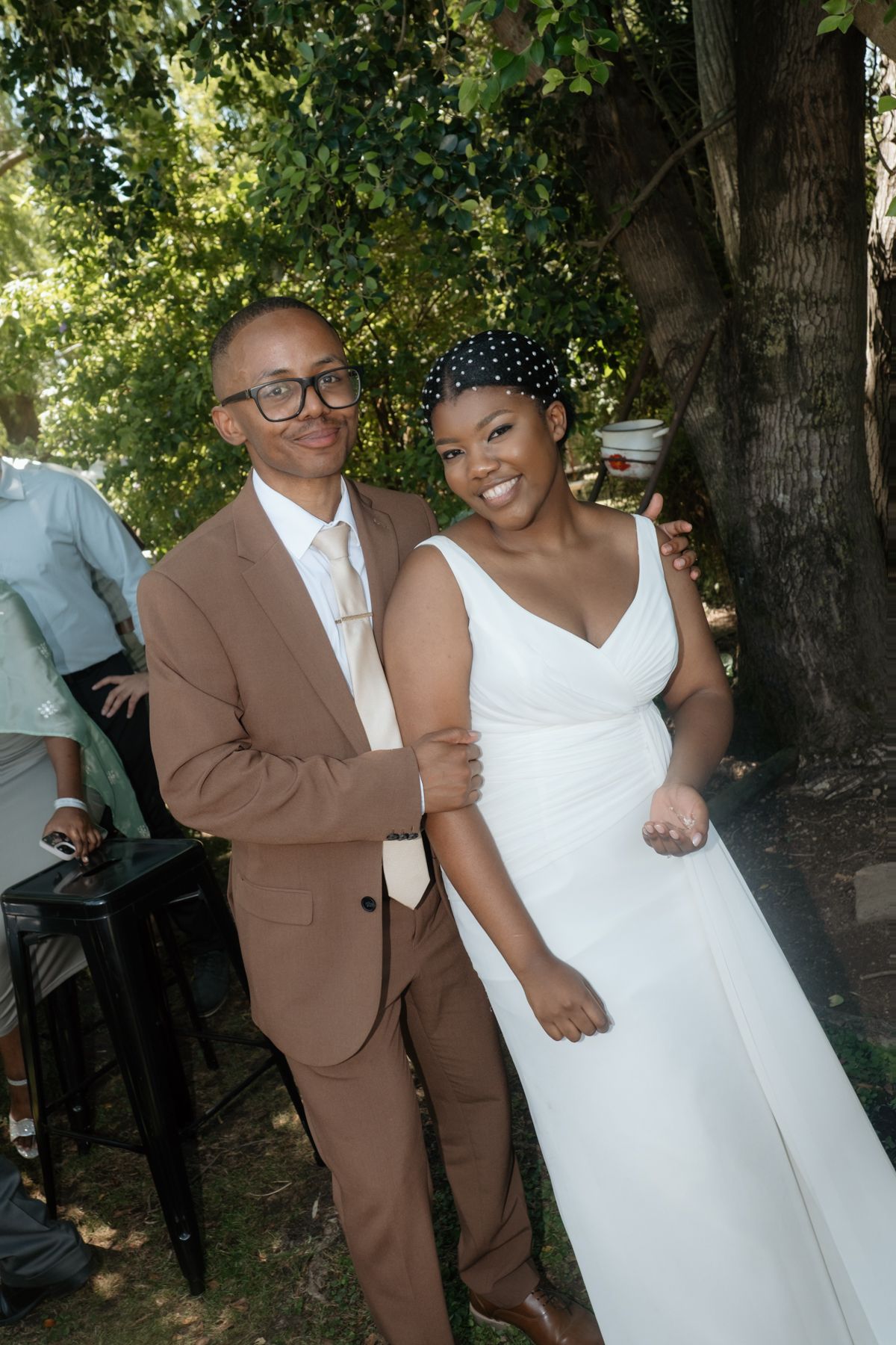 Smiling bride in white gown and groom in tan suit posing together outdoors under trees at their wedding reception.