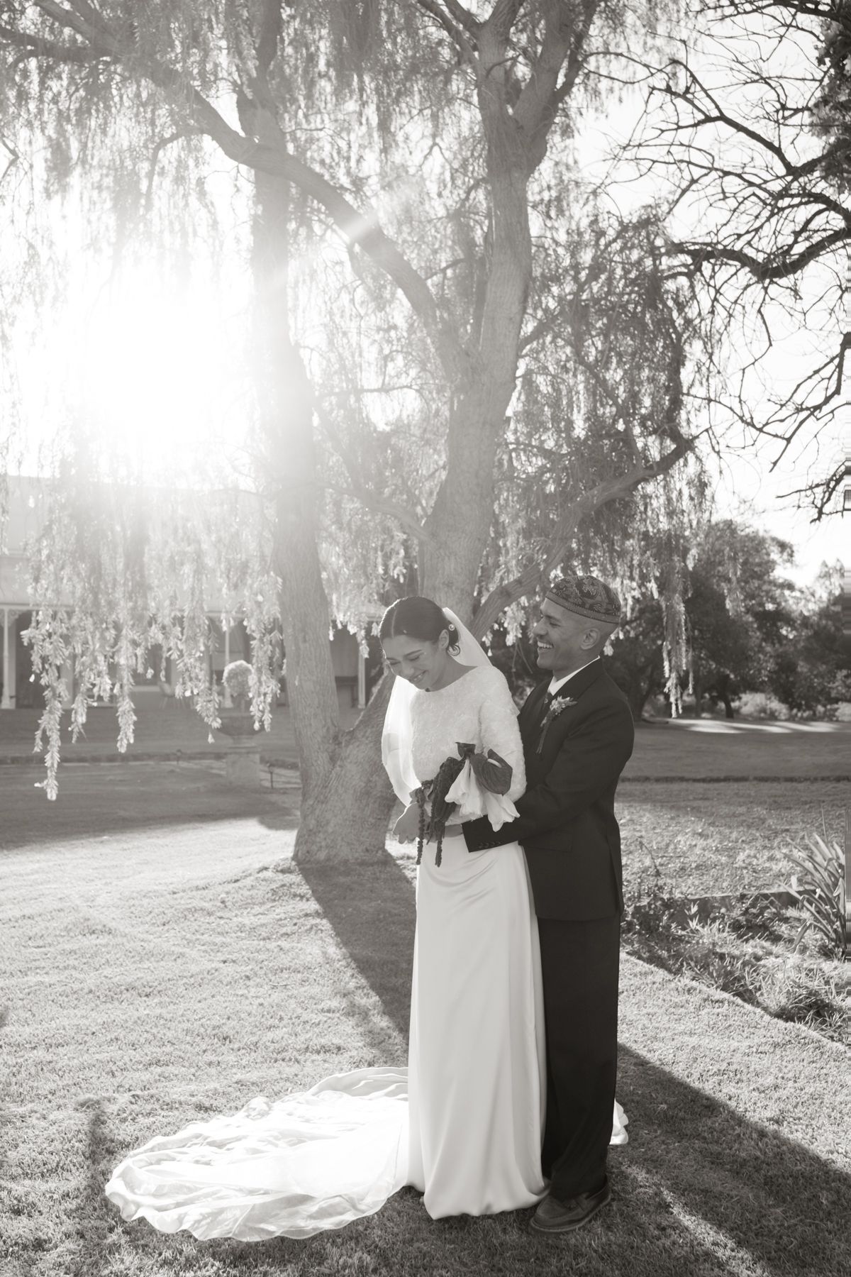 Black and white photo of bride and groom sharing tender moment under sunlit weeping willow tree.