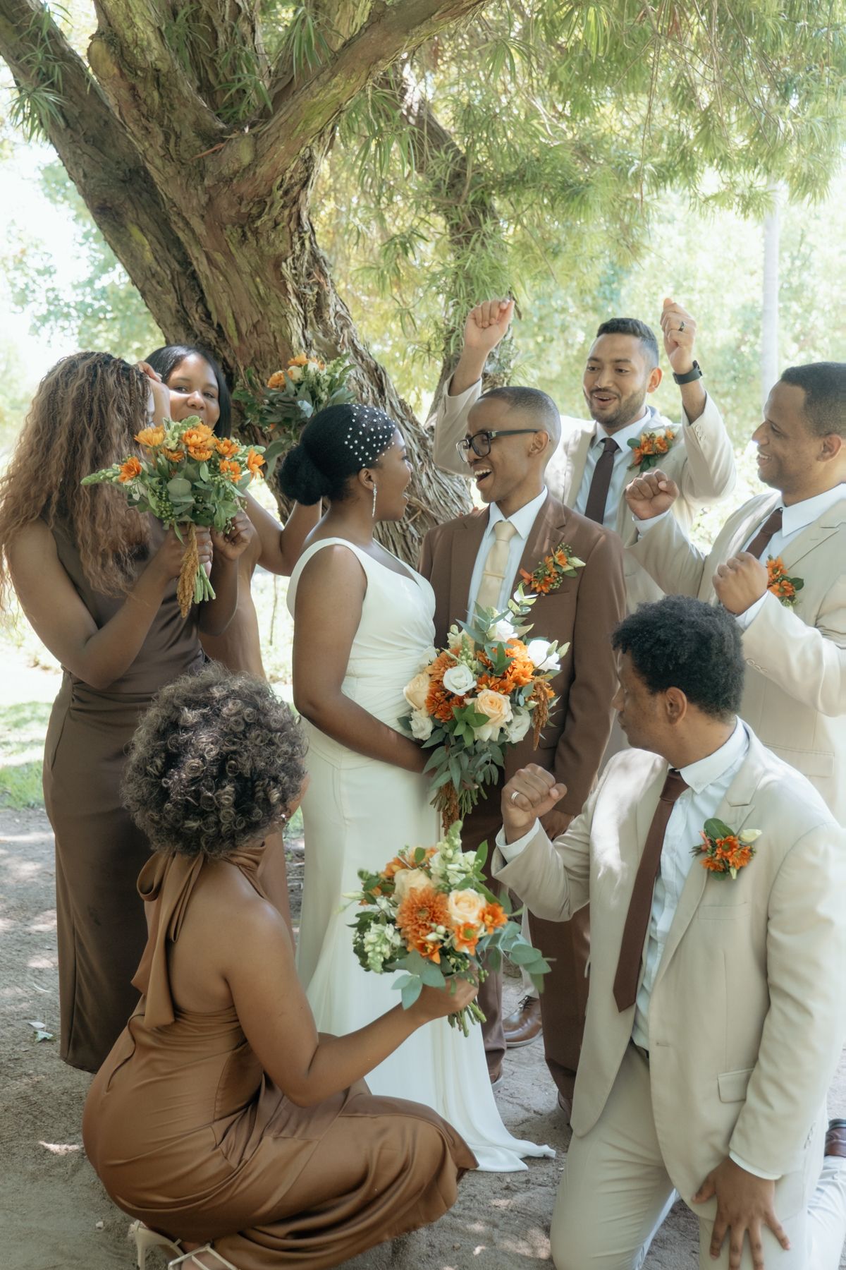 Joyful wedding party celebrates under a tree, bride in white with orange bouquet surrounded by bridesmaids and groomsmen.