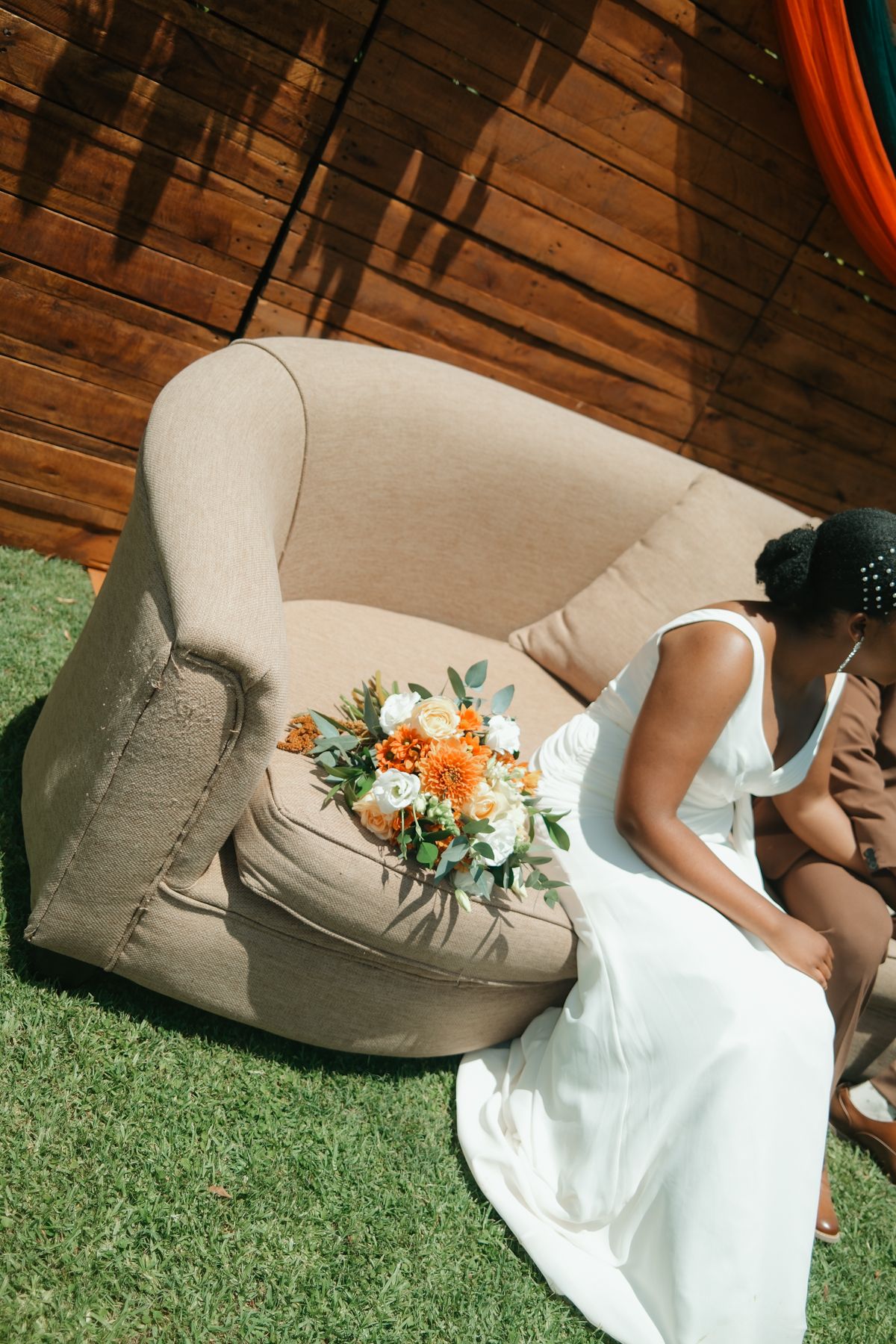 Bride in white dress sits on beige armchair with orange and white bouquet on sunny outdoor wedding day.
