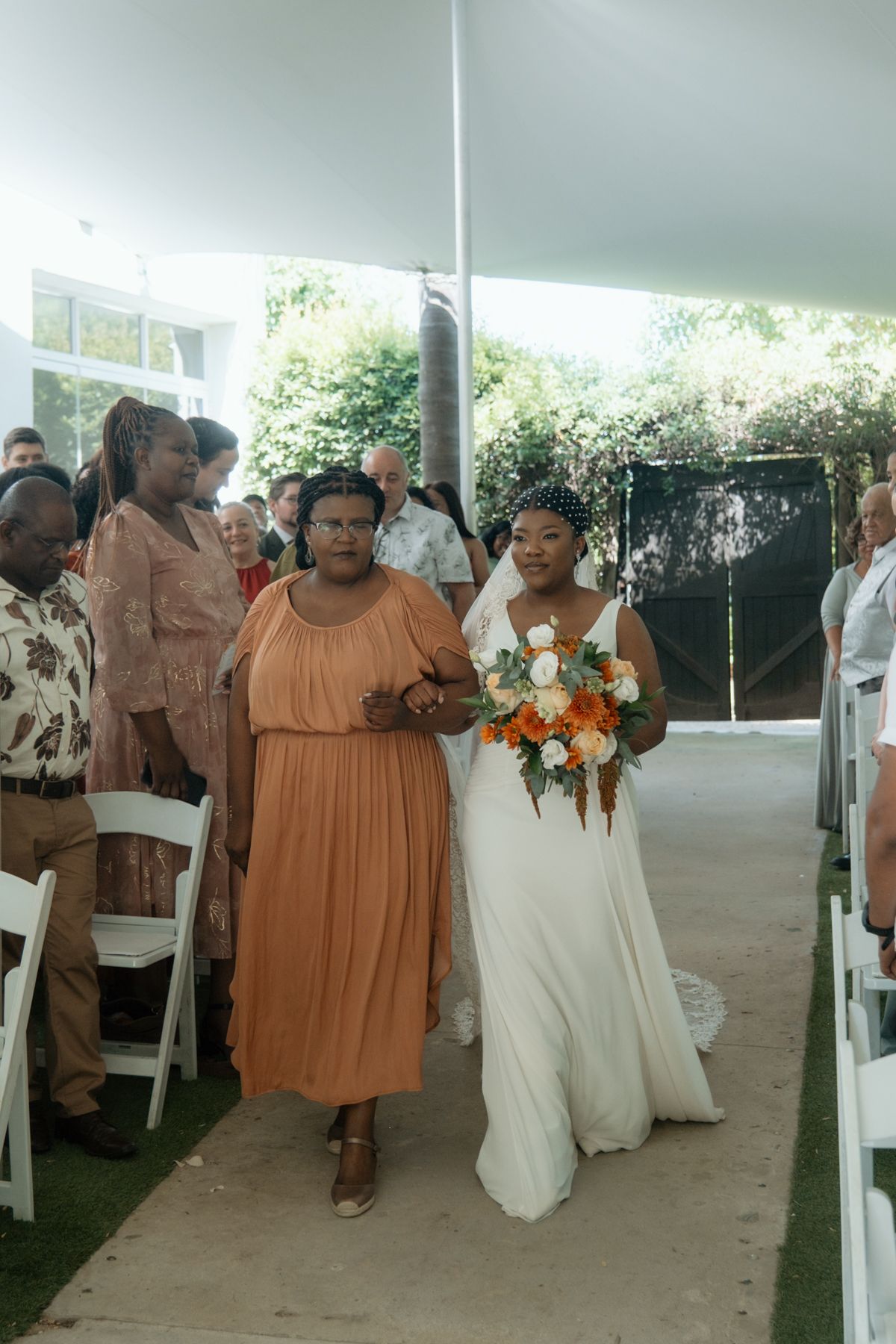 Bride in white gown walks down the aisle with her mother in orange dress at an outdoor tent wedding.