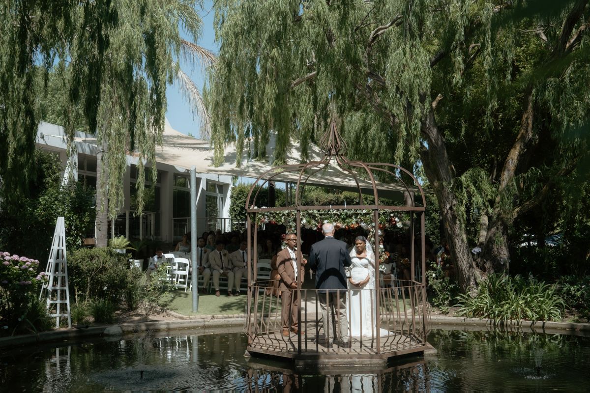 Bride and groom stand in ornate gazebo over pond surrounded by weeping willow trees.