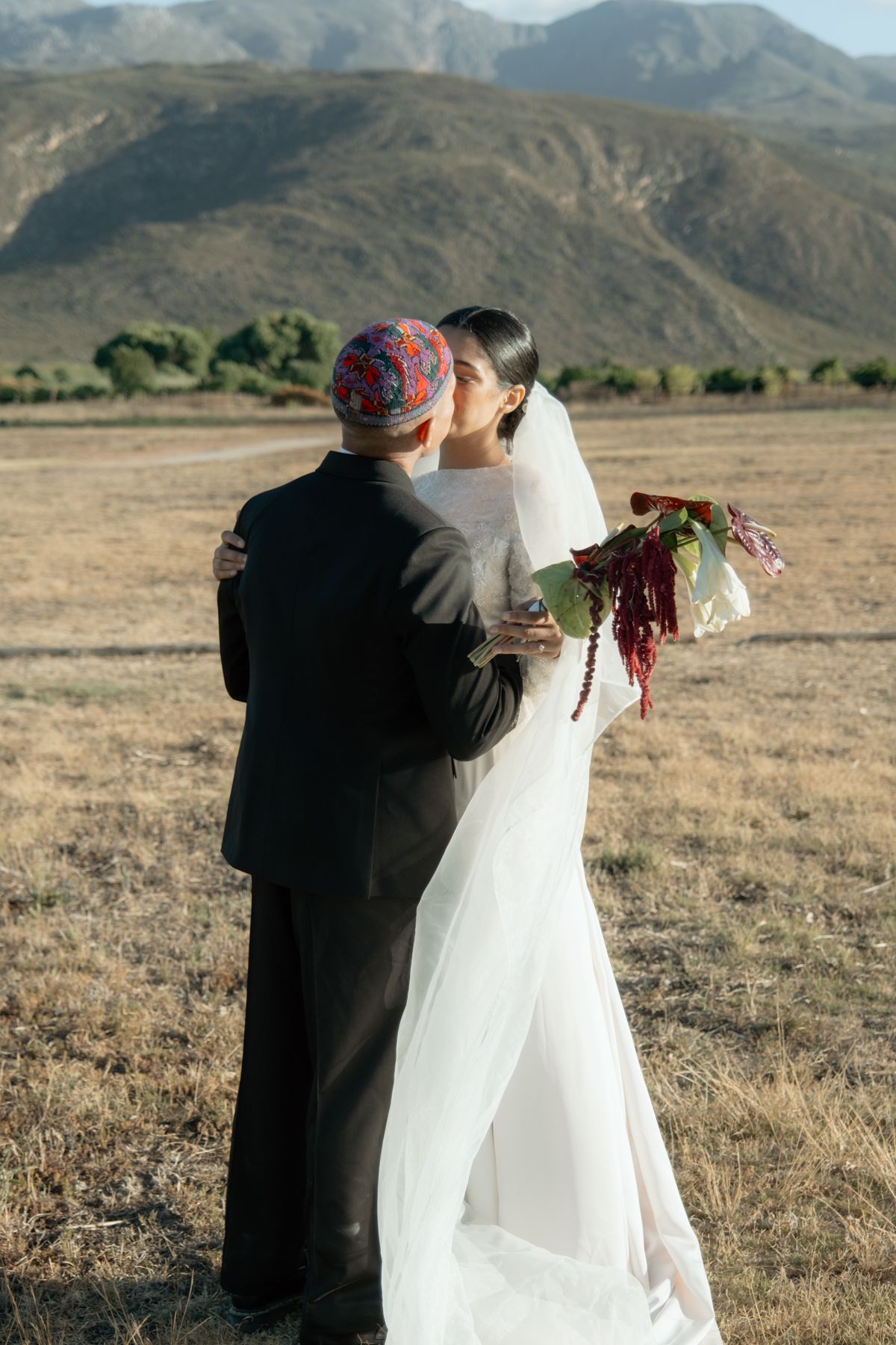 Bride and groom embrace in golden field with stunning mountain landscape at outdoor wedding.