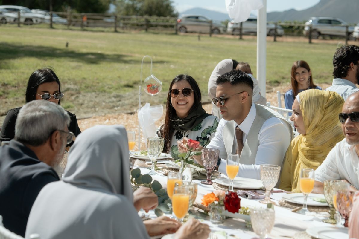 Wedding guests dining outdoors at a table with floral centerpieces and orange juice in sunny field.