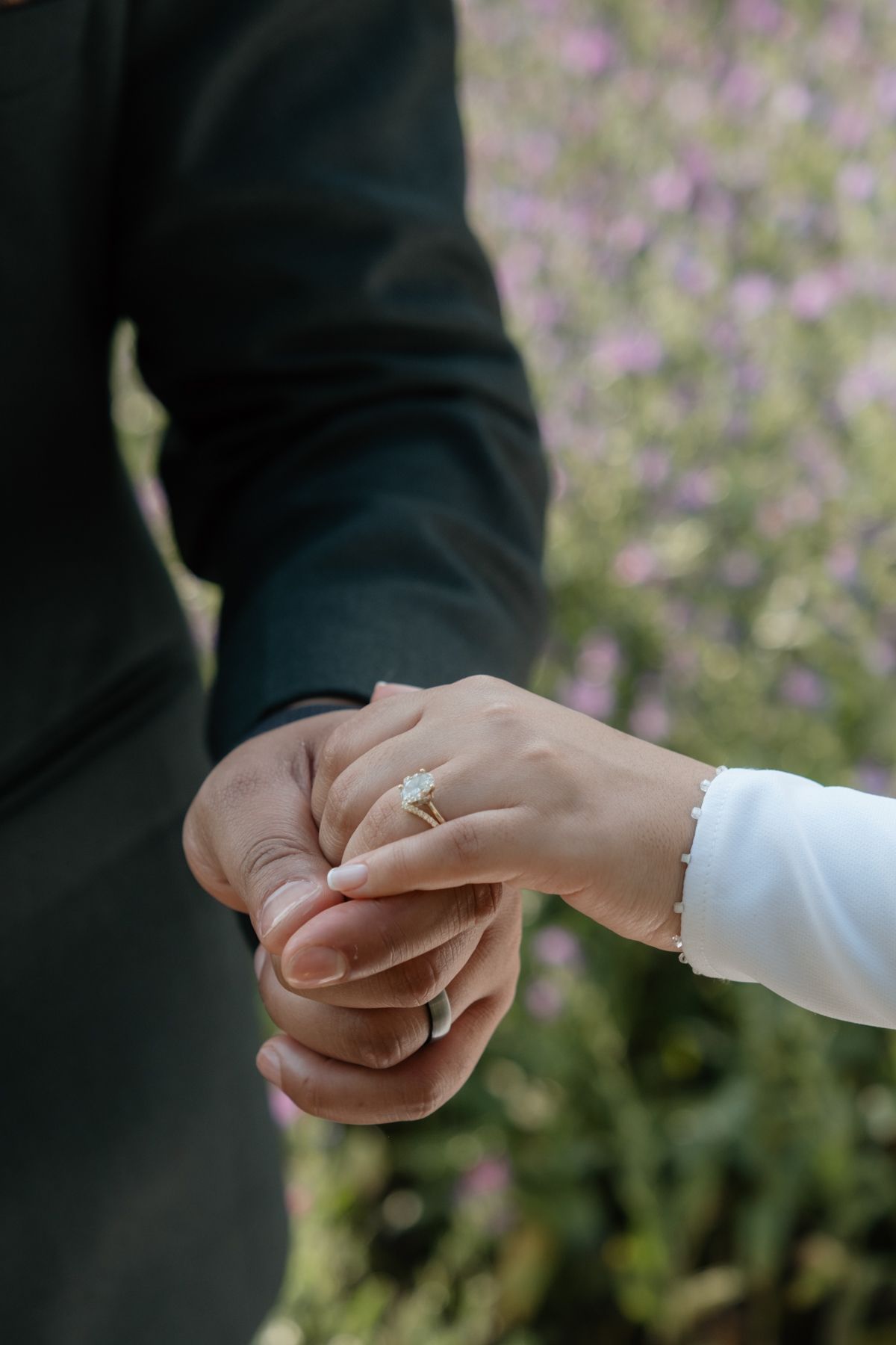 Close-up of bride and groom holding hands with diamond engagement ring in a flower field.