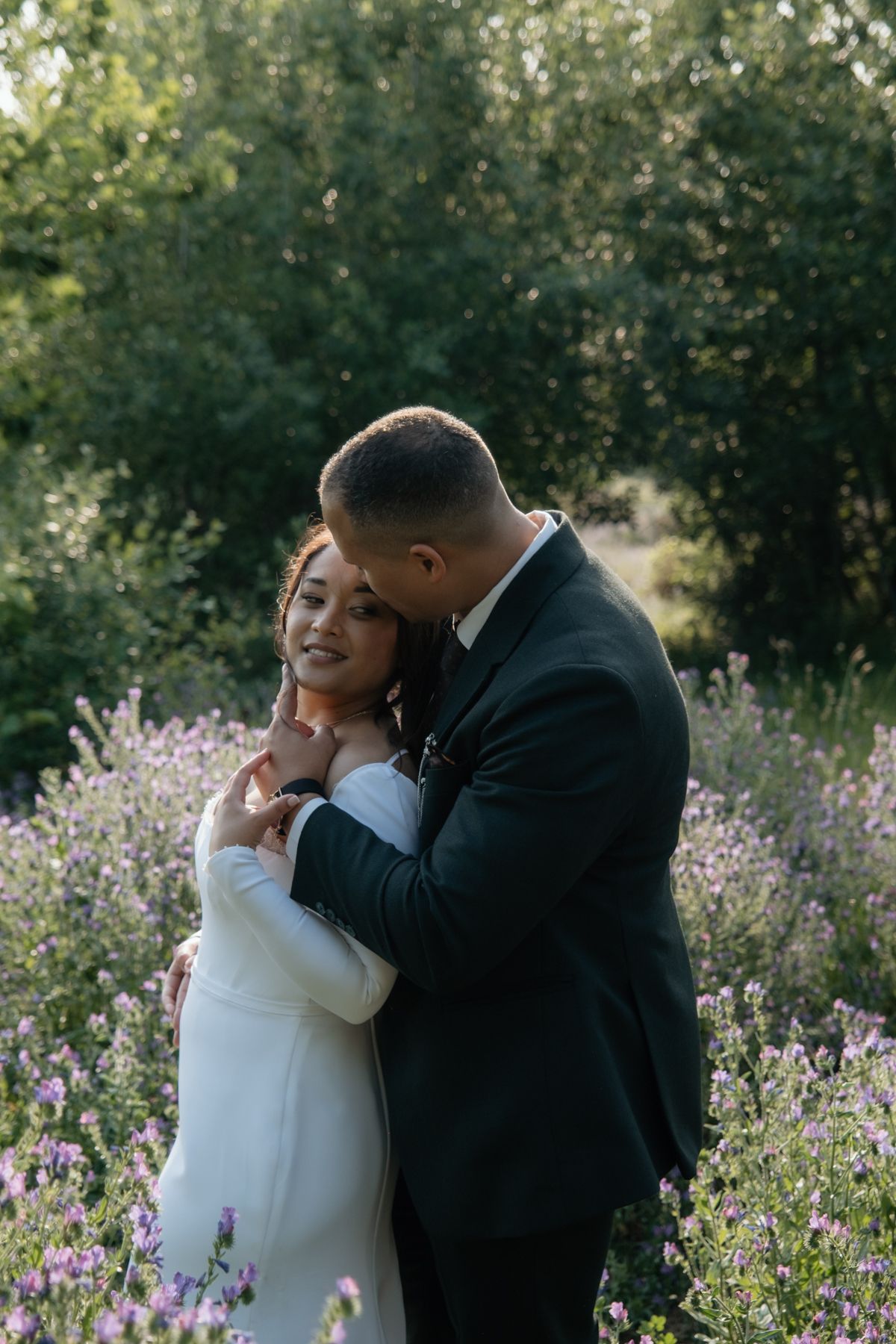 Groom tenderly embraces bride from behind in a lavender field, bride smiling in white long-sleeved wedding gown.