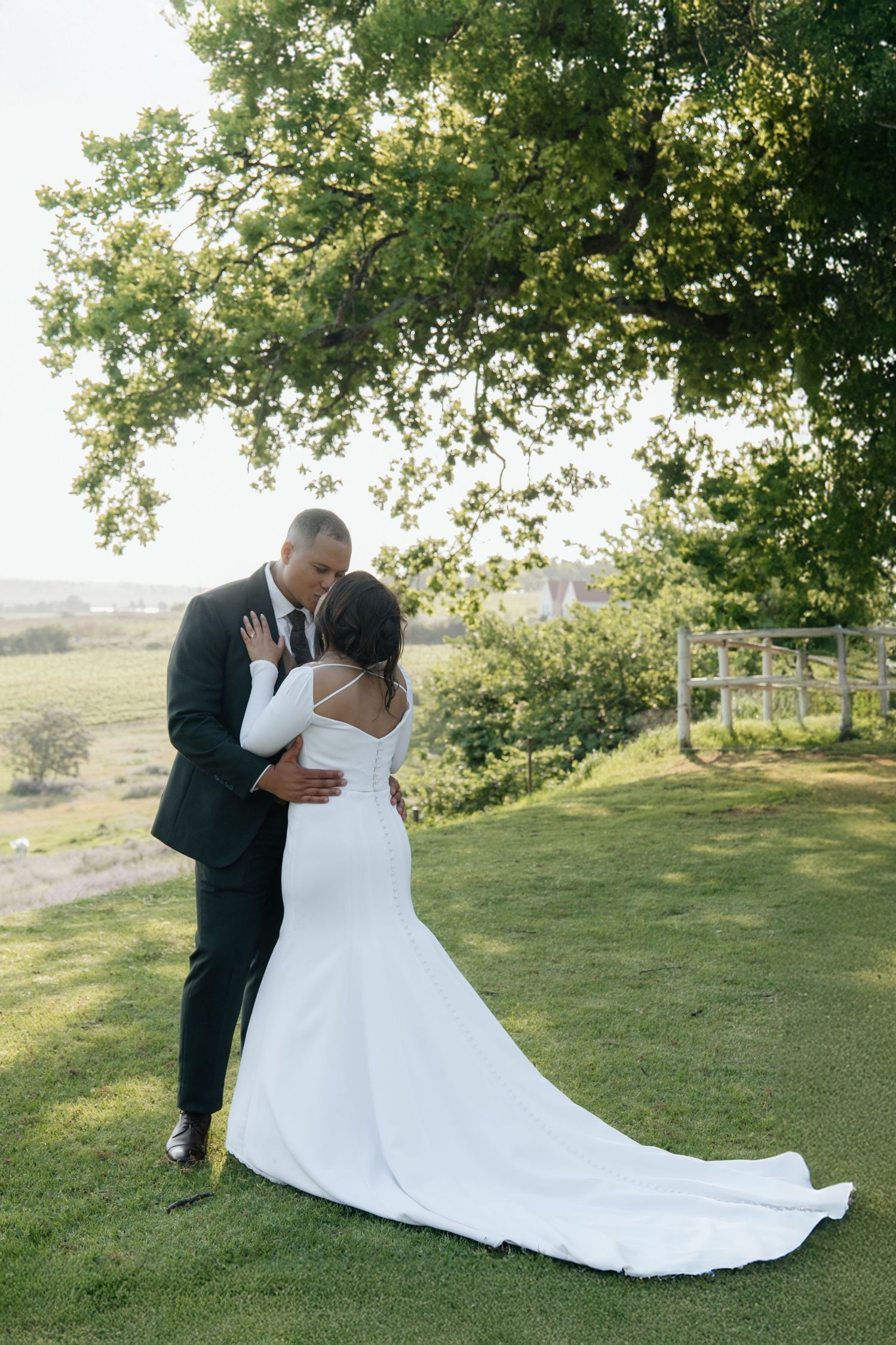Bride and groom share romantic kiss under large tree in lush garden, bride in fitted white gown with train.