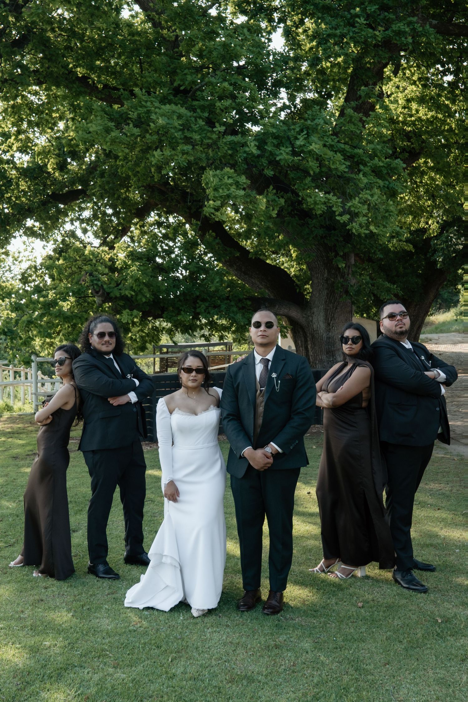 Bride and groom with wedding party wearing sunglasses striking a serious pose under a green tree.