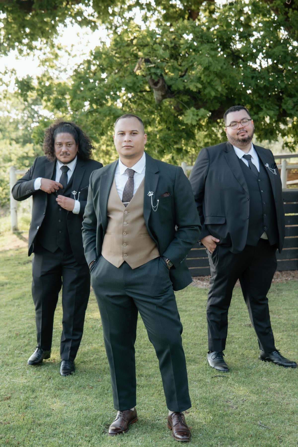 Three groomsmen in dark suits and tan vests pose confidently outdoors under a large green tree at a wedding.