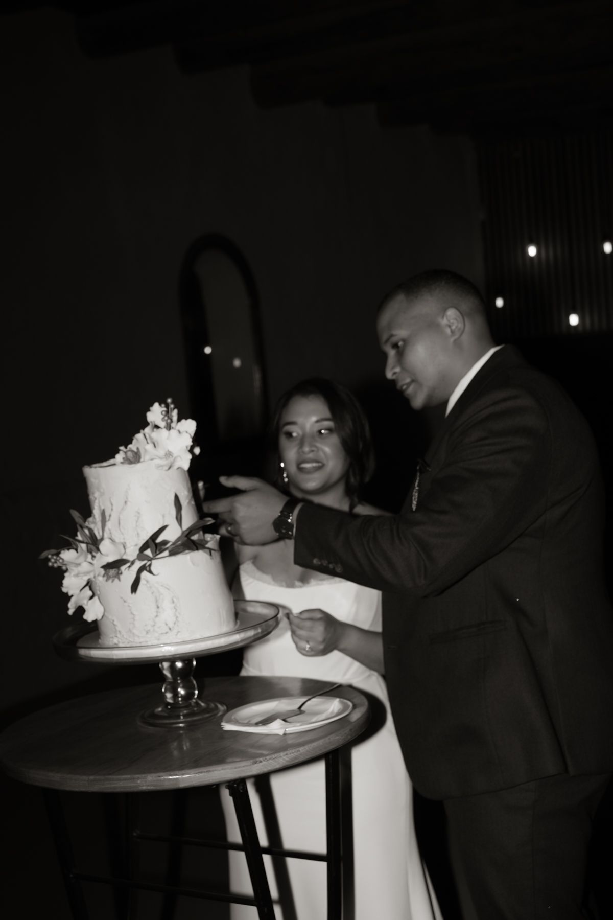 Black and white photo of couple cutting elegant two-tier floral wedding cake at dark romantic reception.