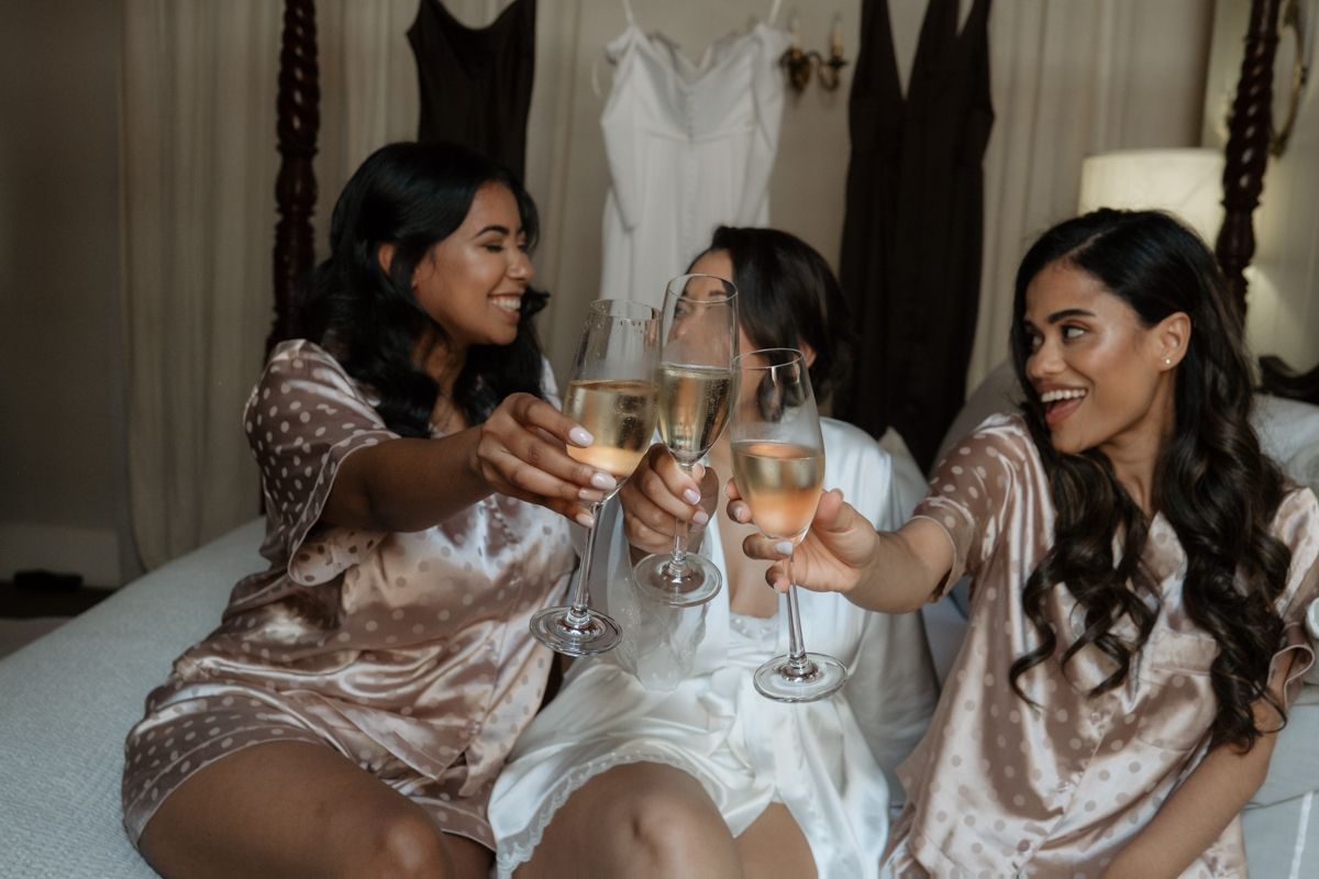 Three bridesmaids in satin robes toasting champagne glasses while sitting on bed before wedding.