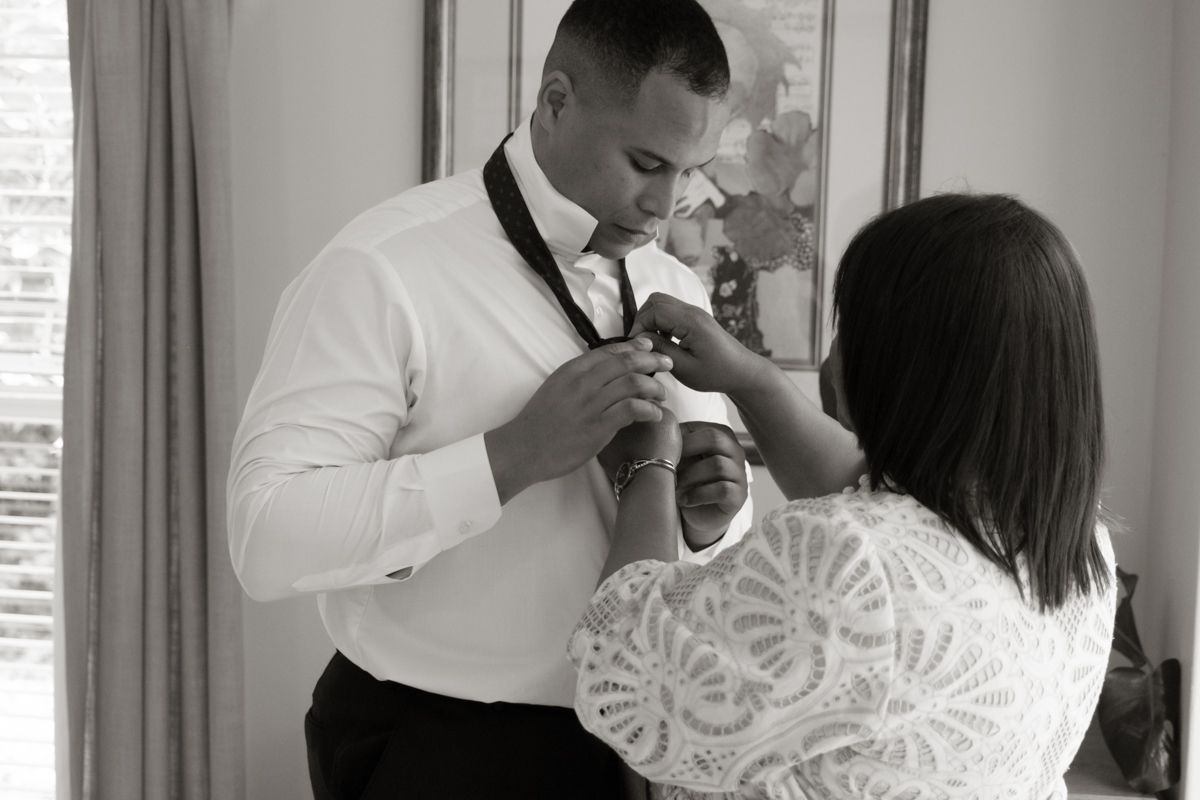 Groom in white shirt gets bow tie adjusted by woman in lace top, elegant black and white wedding photo.