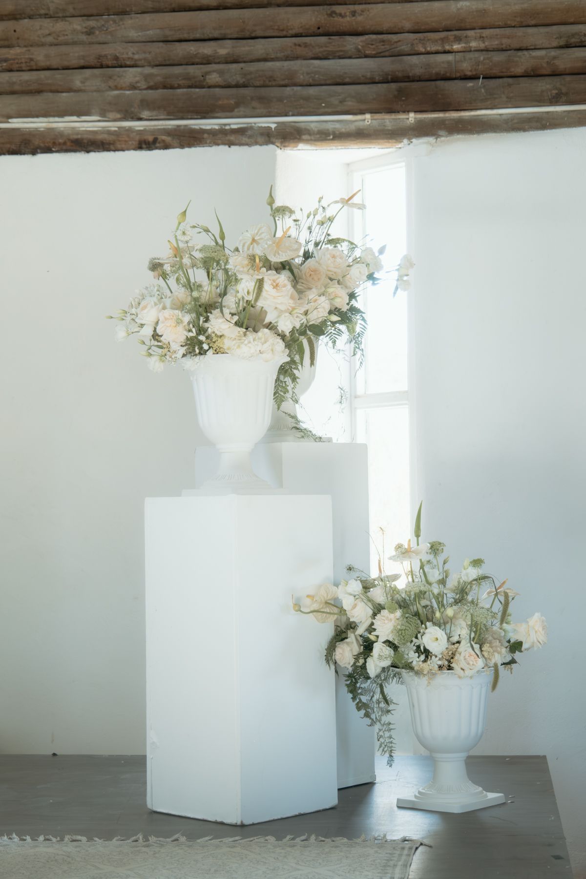 Two white urn vases with elegant white floral arrangements displayed on white pedestals in a bright minimalist wedding venue.