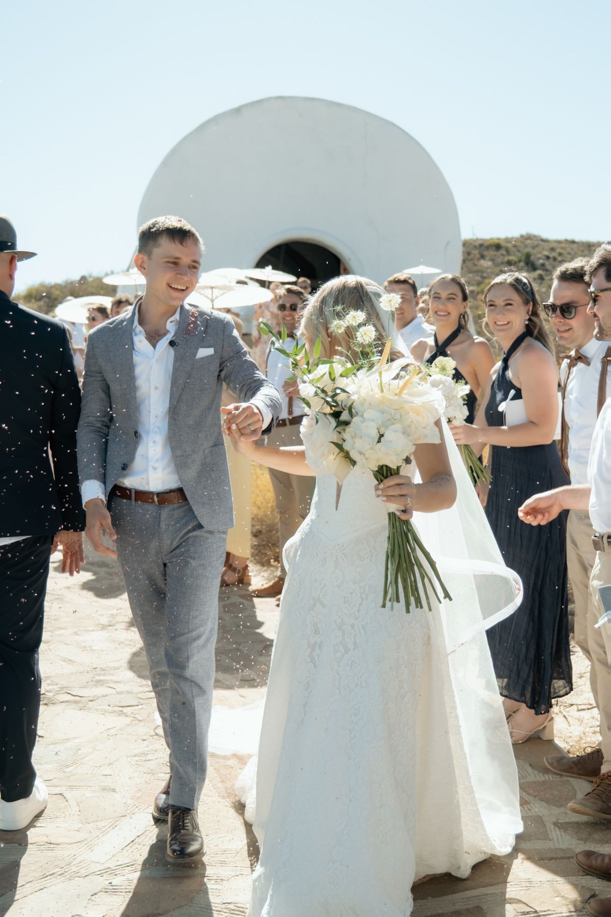 Newlyweds walking hand in hand after outdoor wedding ceremony as guests celebrate around them.