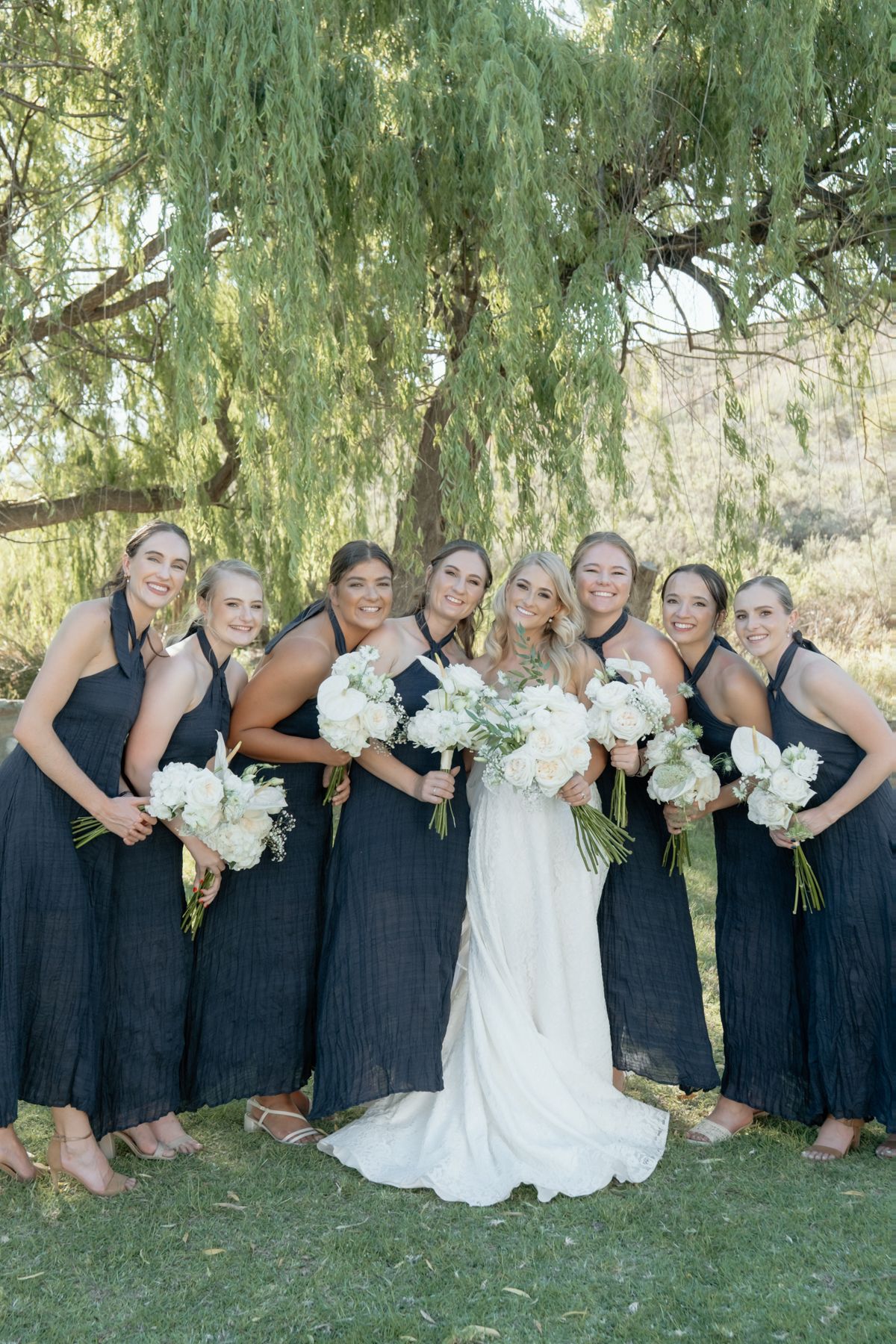 Bride in white gown poses with six bridesmaids in navy dresses holding white bouquets under willow tree.
