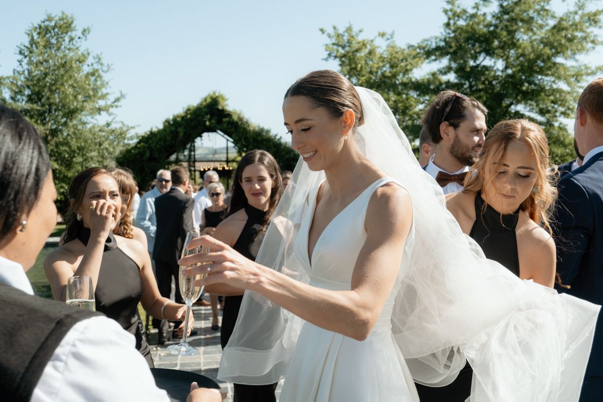 Smiling bride in a white gown pours champagne while mingling with guests at an outdoor wedding reception.