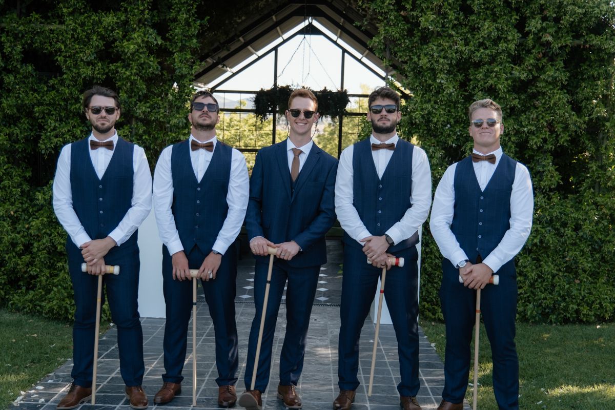 Five groomsmen in matching navy vests and bow ties pose with canes in front of a glass pavilion.