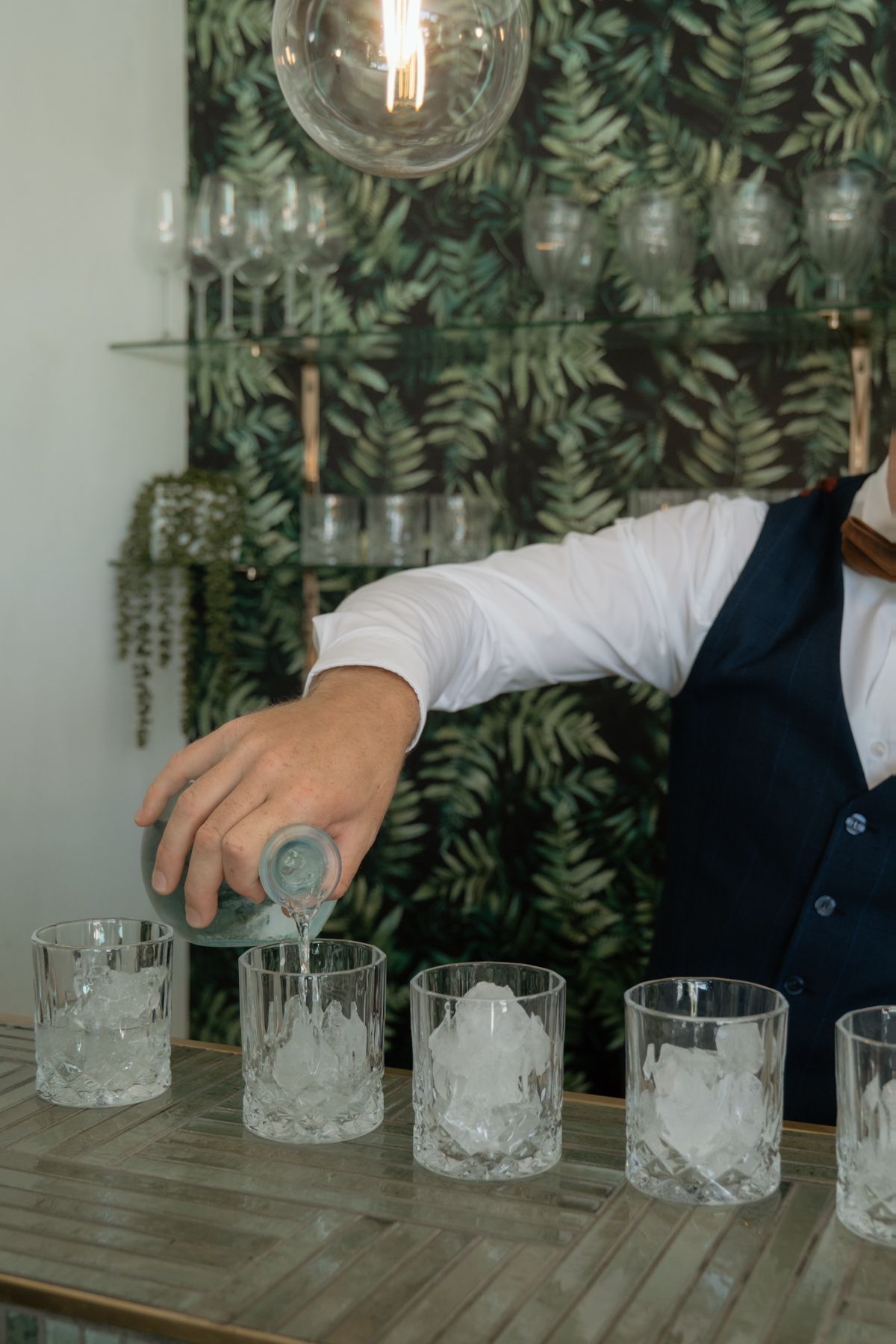 Bartender in navy vest pouring drinks into crystal glasses filled with ice at an elegant bar.