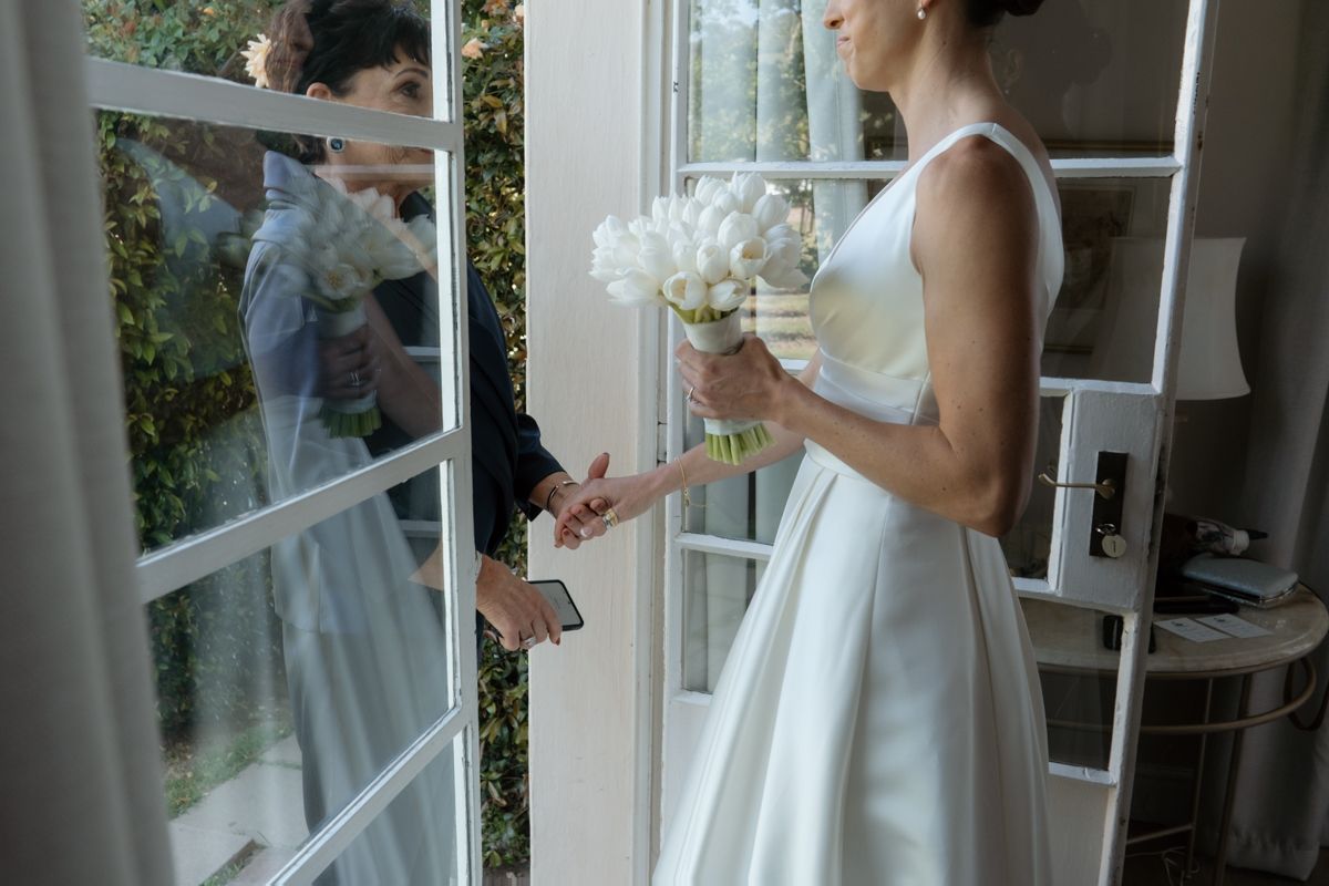 Bride in white dress holds white bouquet while exchanging rings at window with guest.