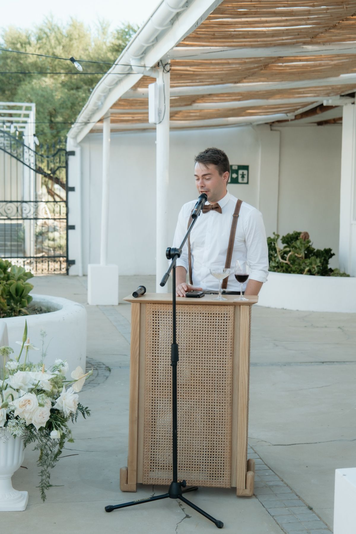 Man in white shirt and brown bow tie delivering a speech at a wooden podium during a wedding reception.