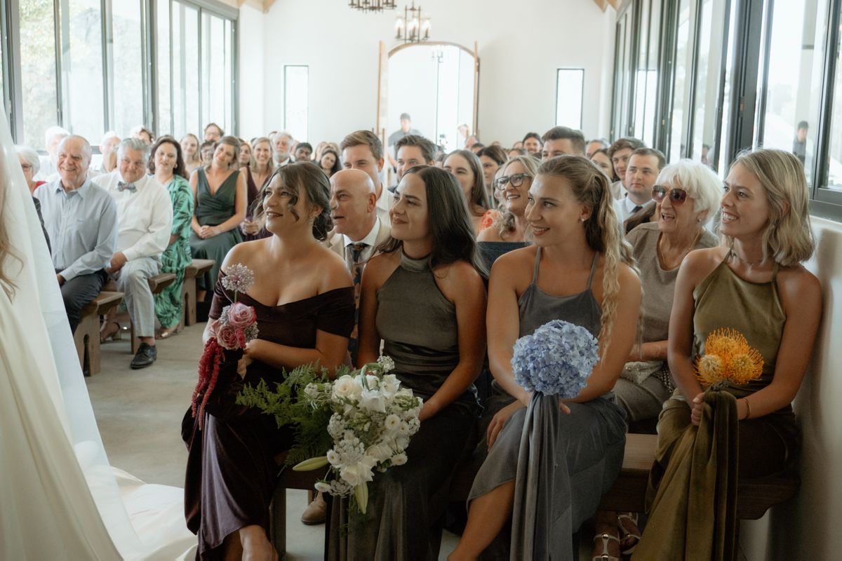 Bridesmaids in mismatched gray and burgundy dresses holding bouquets smile during an elegant indoor wedding ceremony.