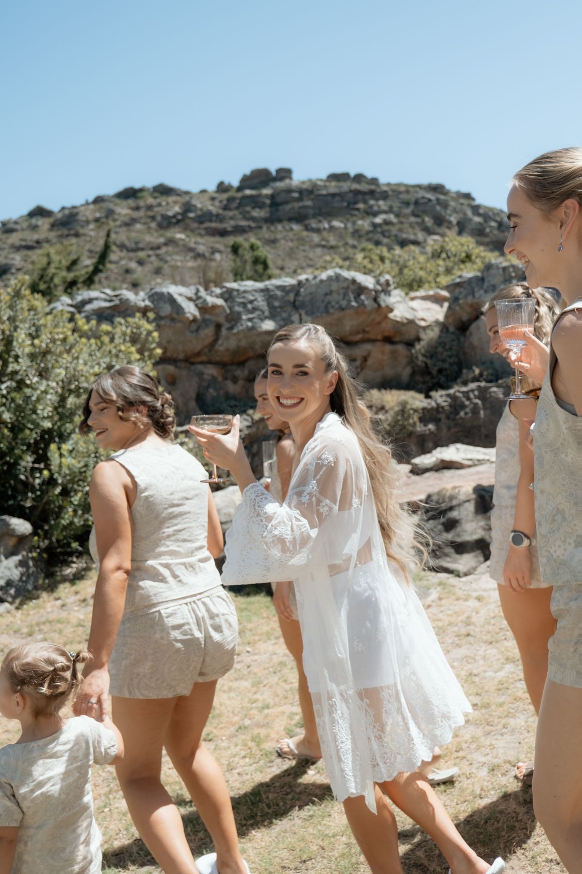Bridesmaids in white and cream outfits celebrating outdoors with champagne against a rocky hillside backdrop.