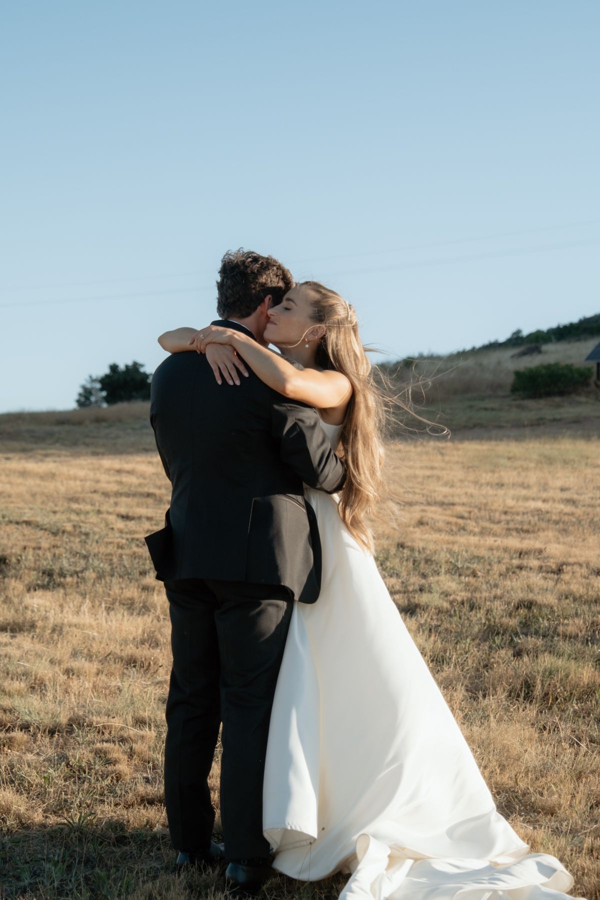 Bride and groom share romantic kiss in golden field under clear blue sky during outdoor wedding.