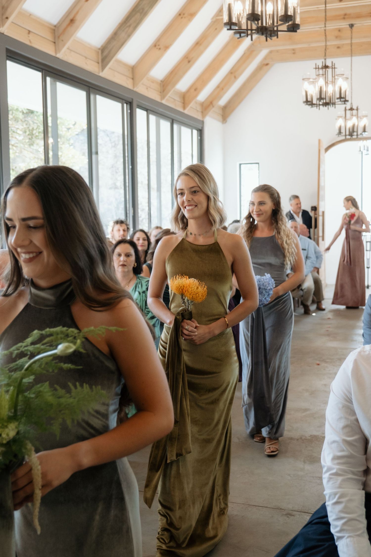 Bridesmaids in olive and grey velvet gowns walk down aisle carrying bouquets in bright barn venue.