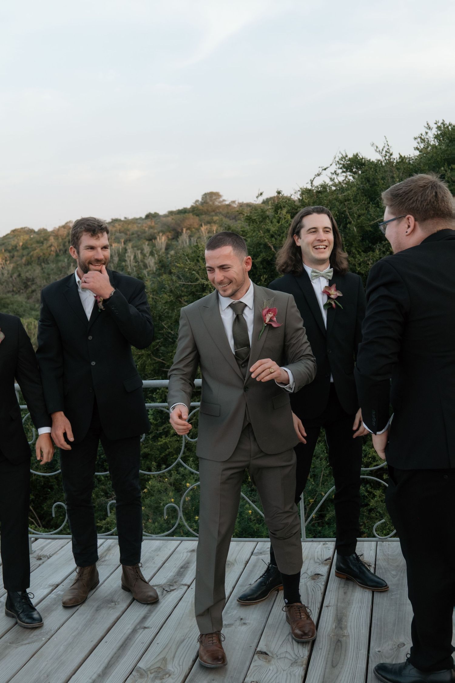 Groom in gray suit laughs with groomsmen in black suits on wooden deck with lush green trees backdrop.
