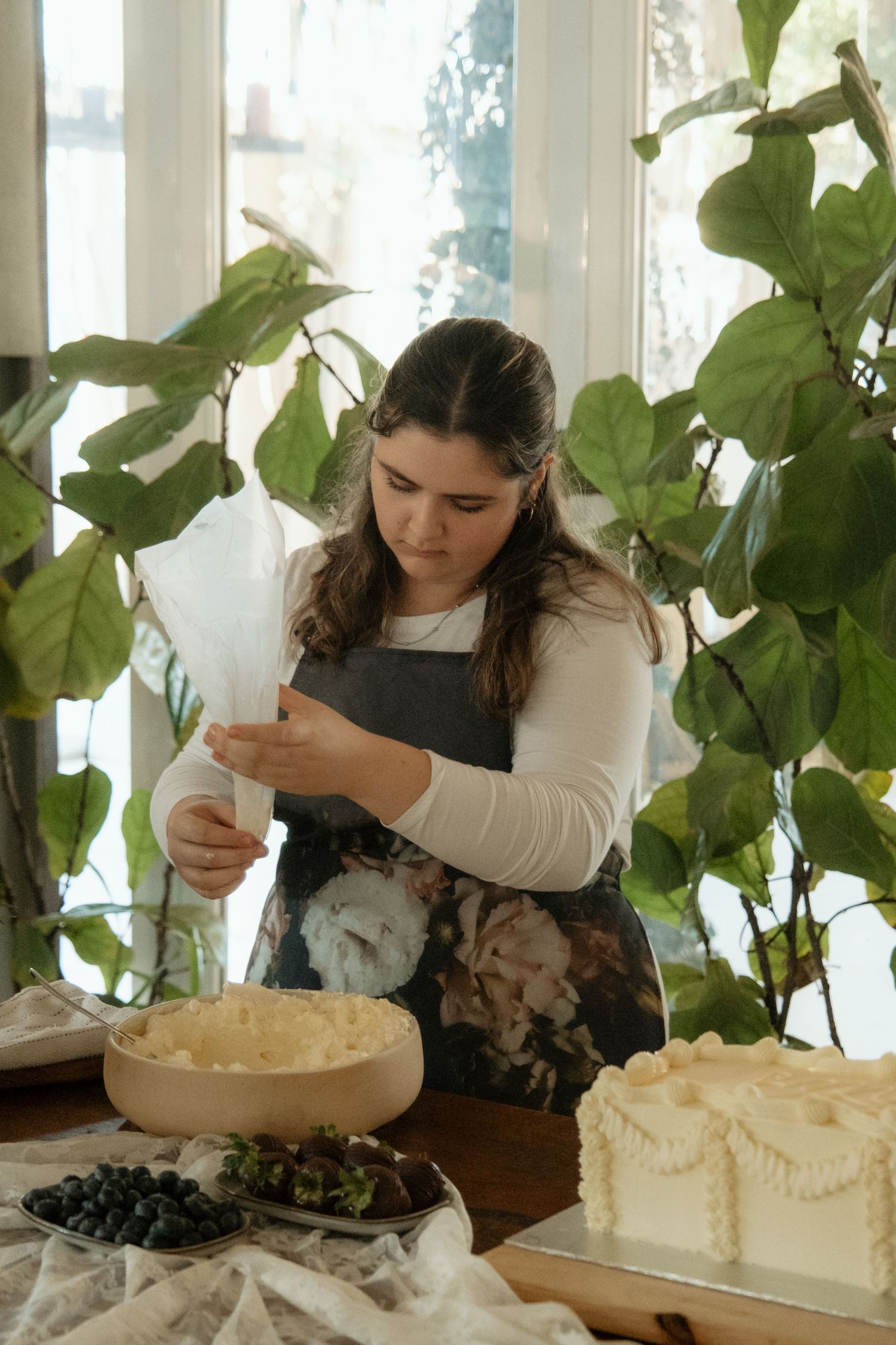 Baker decorating a white wedding cake with piping bag surrounded by blueberries and plants.