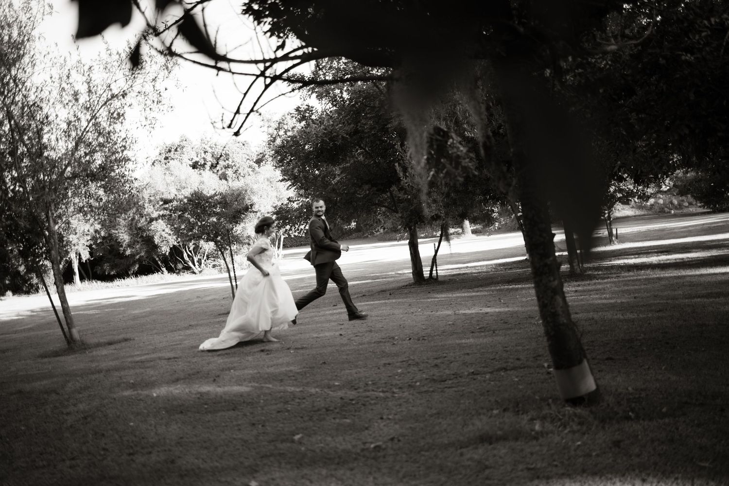 Black and white photo of bride and groom playfully running across a grassy park after their wedding.