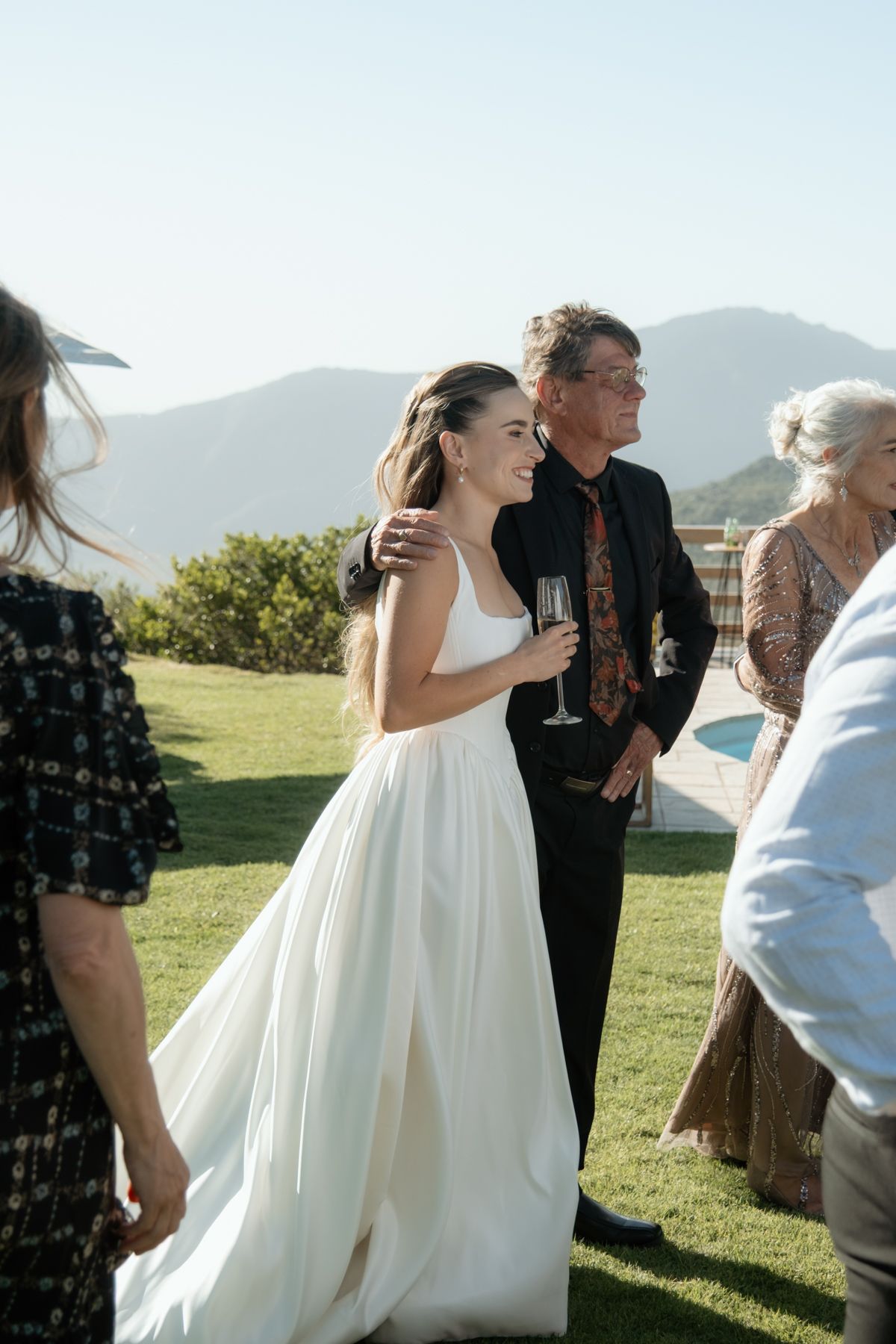 Bride in white gown holding champagne glass with guests and mountain backdrop at outdoor wedding.