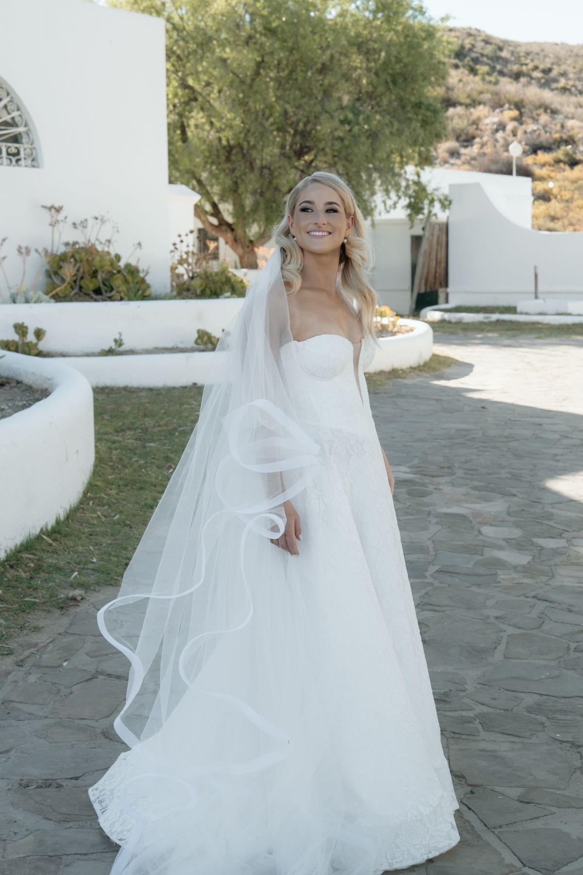 Radiant bride in a strapless ball gown with flowing cathedral veil posing outdoors at a white Mediterranean-style wedding venue.