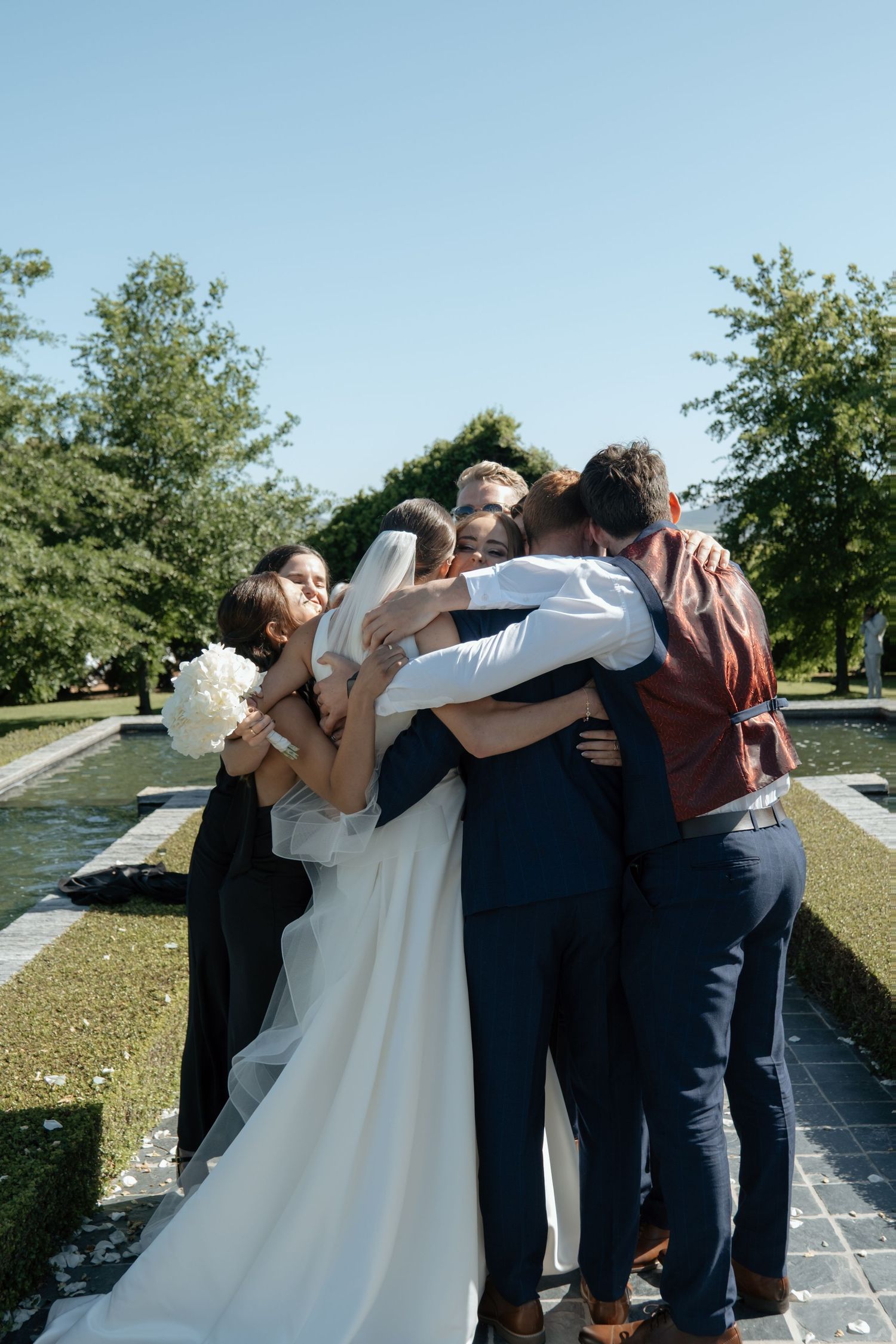 Wedding party sharing a group hug outdoors on sunny wedding day.