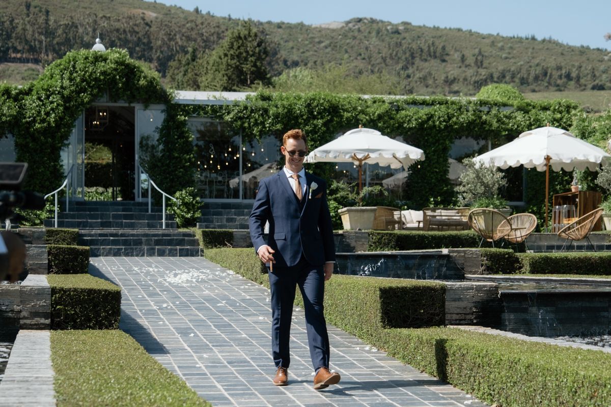 Groom in a navy blue suit standing on a garden pathway at an elegant vineyard wedding venue.