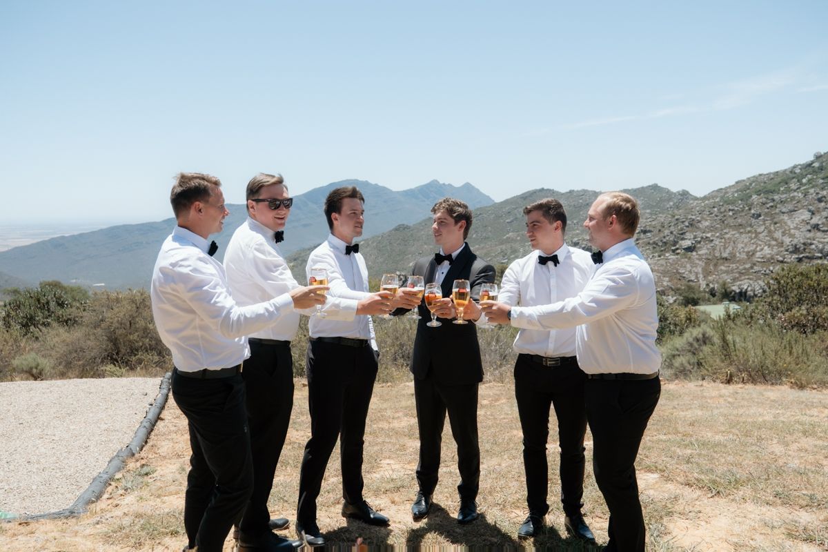 Groomsmen in black tuxedos toasting with drinks on a sunny mountain backdrop at a wedding celebration.