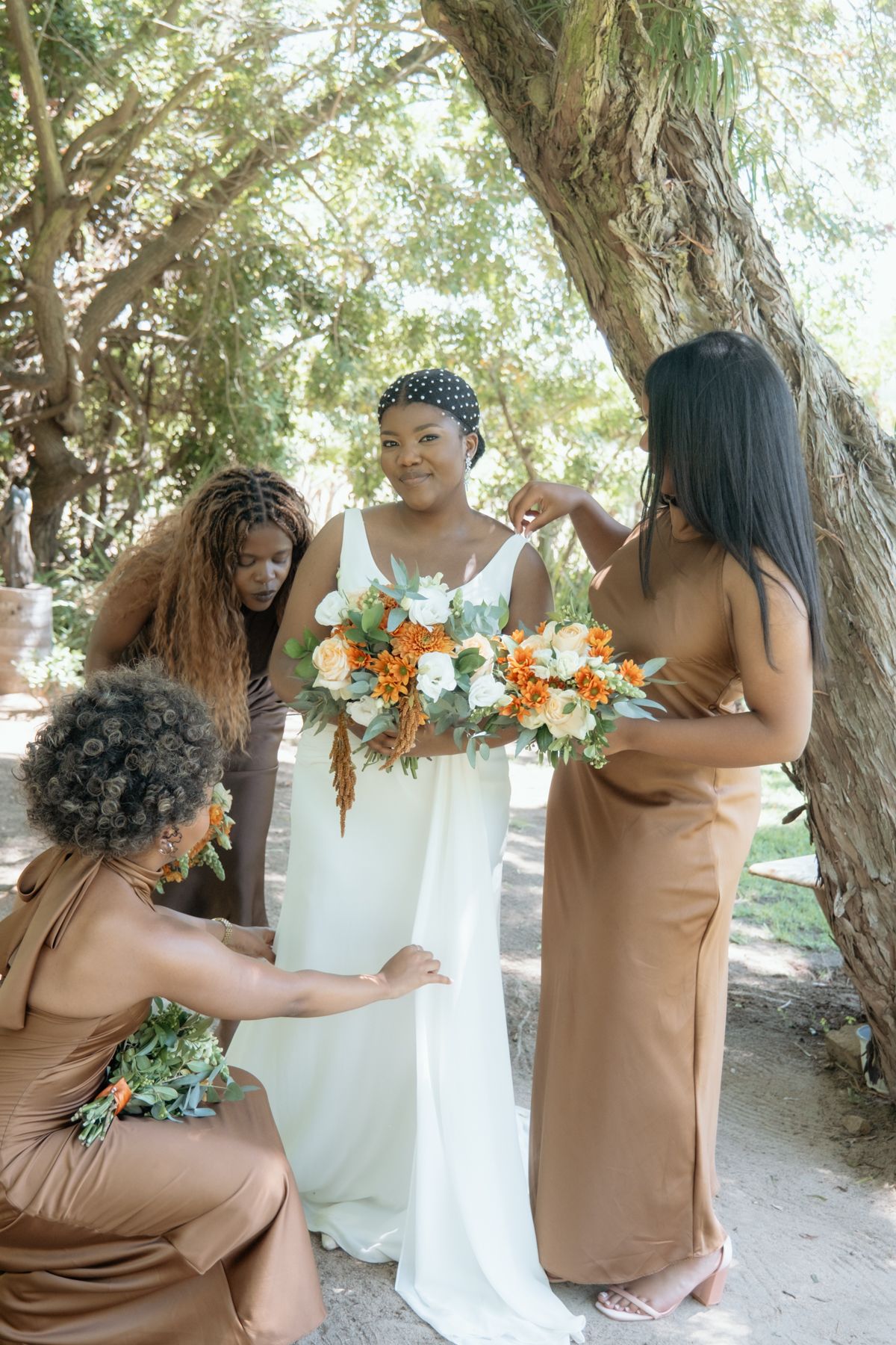 Bridesmaids in brown satin dresses help adjust bride in white gown while holding orange and white bouquets outdoors.