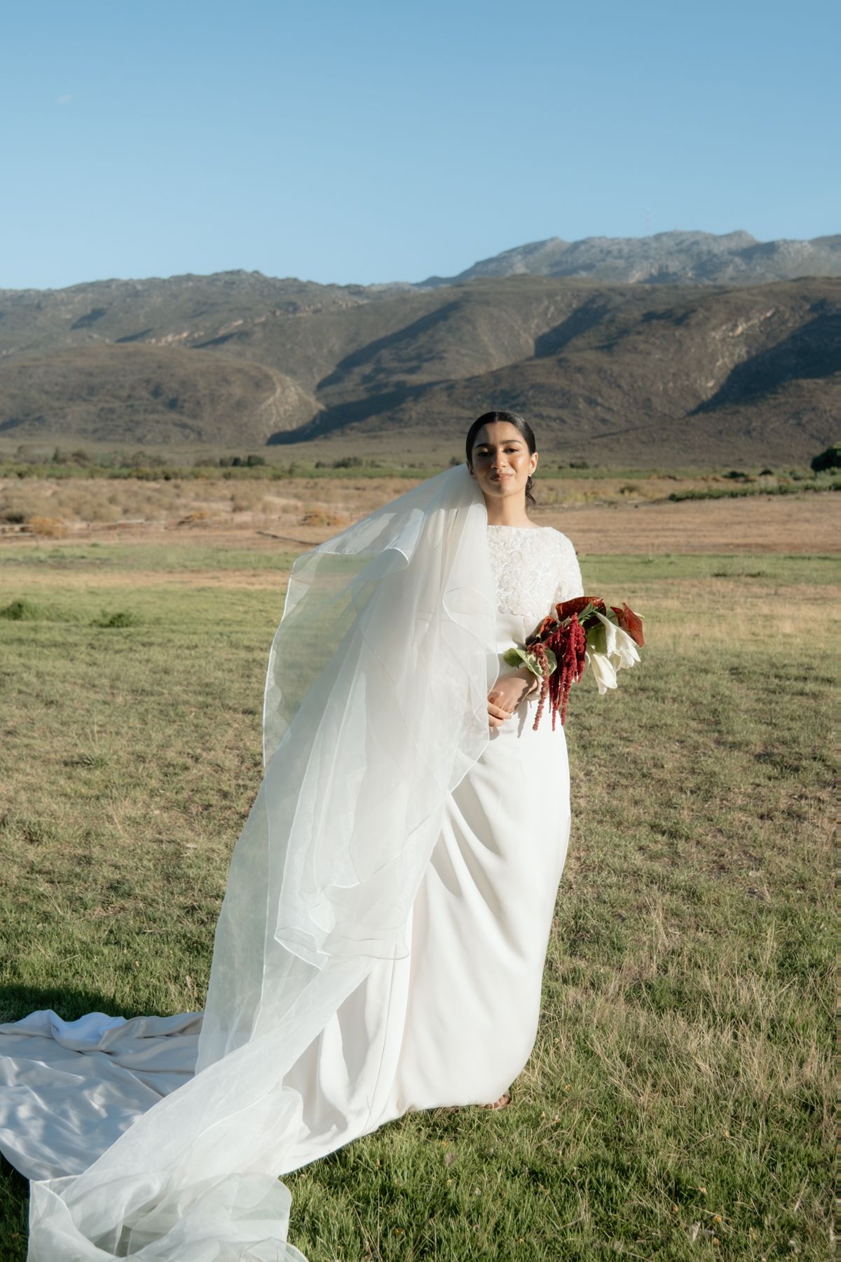 Elegant bride in white gown and veil holding a red and white bouquet with scenic mountain landscape behind her.