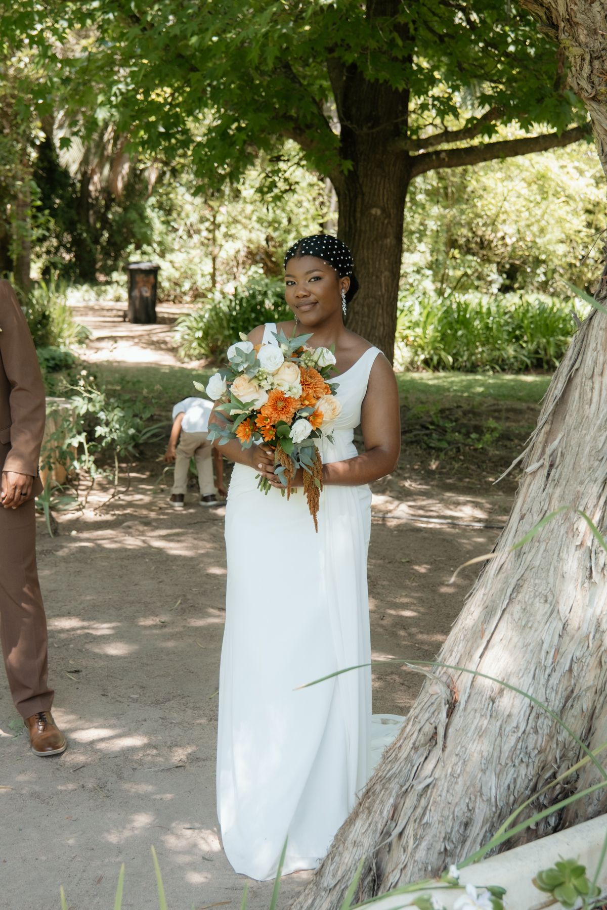 A smiling Black bride in a white gown holds a vibrant orange and white bouquet while walking through a lush garden setting.