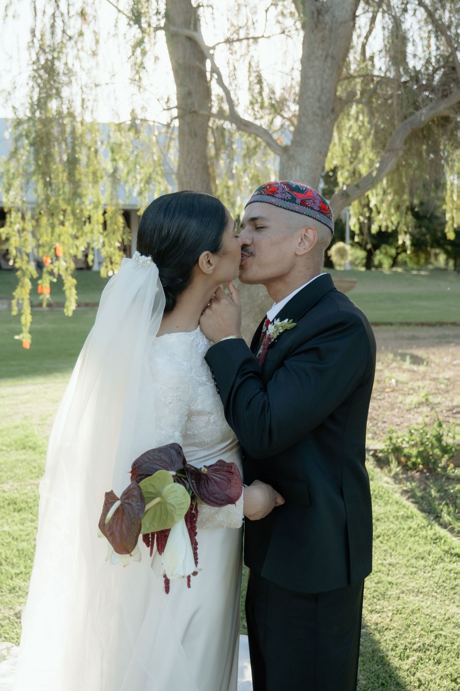 Bride and groom share a kiss outdoors, bride holding a dark floral bouquet, groom wearing a traditional cap.