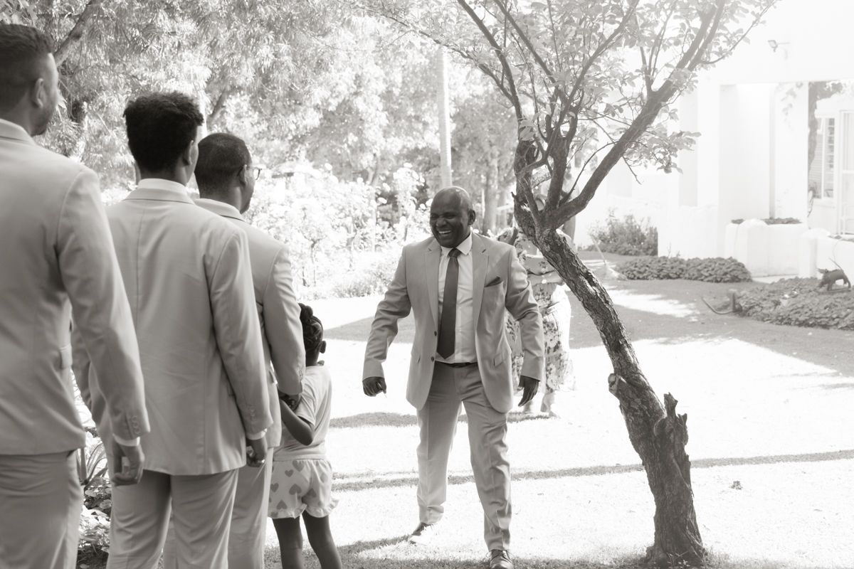 Black and white photo of groomsmen in suits gathered outdoors near trees before a wedding ceremony.